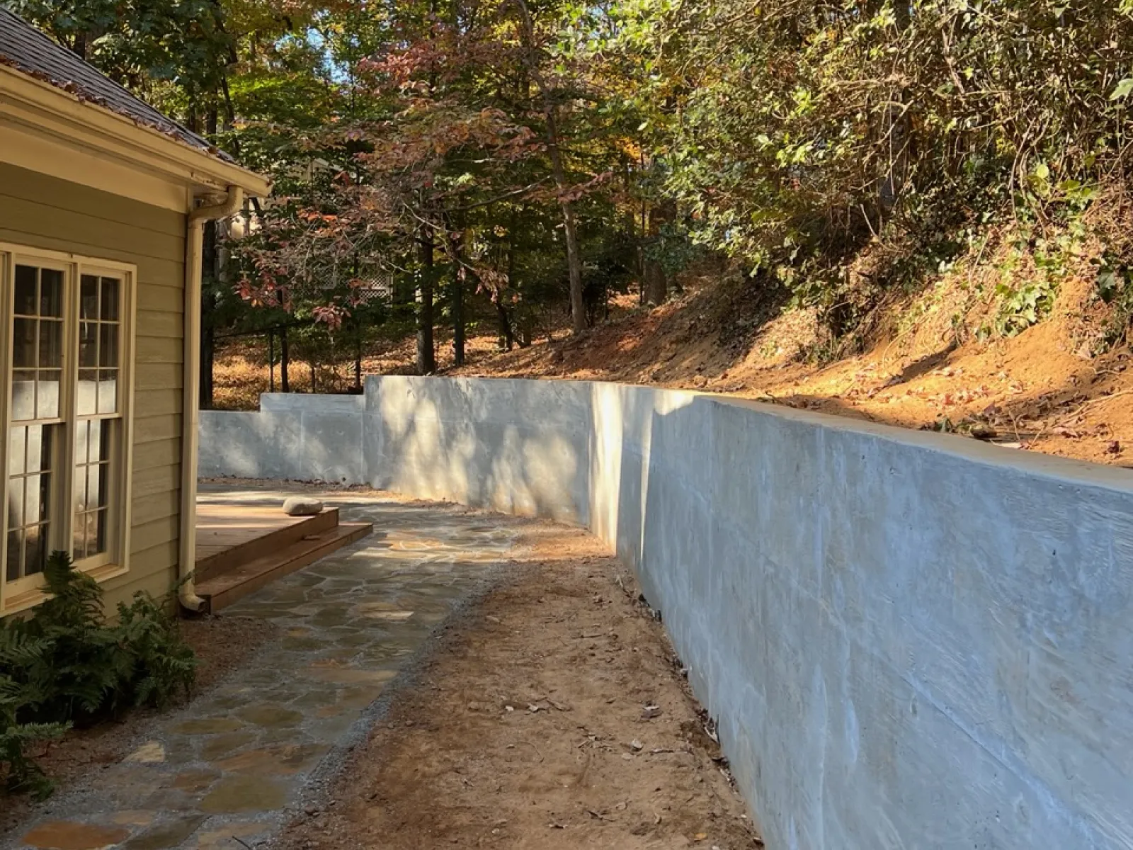 A serene backyard view featuring a stone path beside a newly constructed concrete wall and surrounded by autumn foliage.