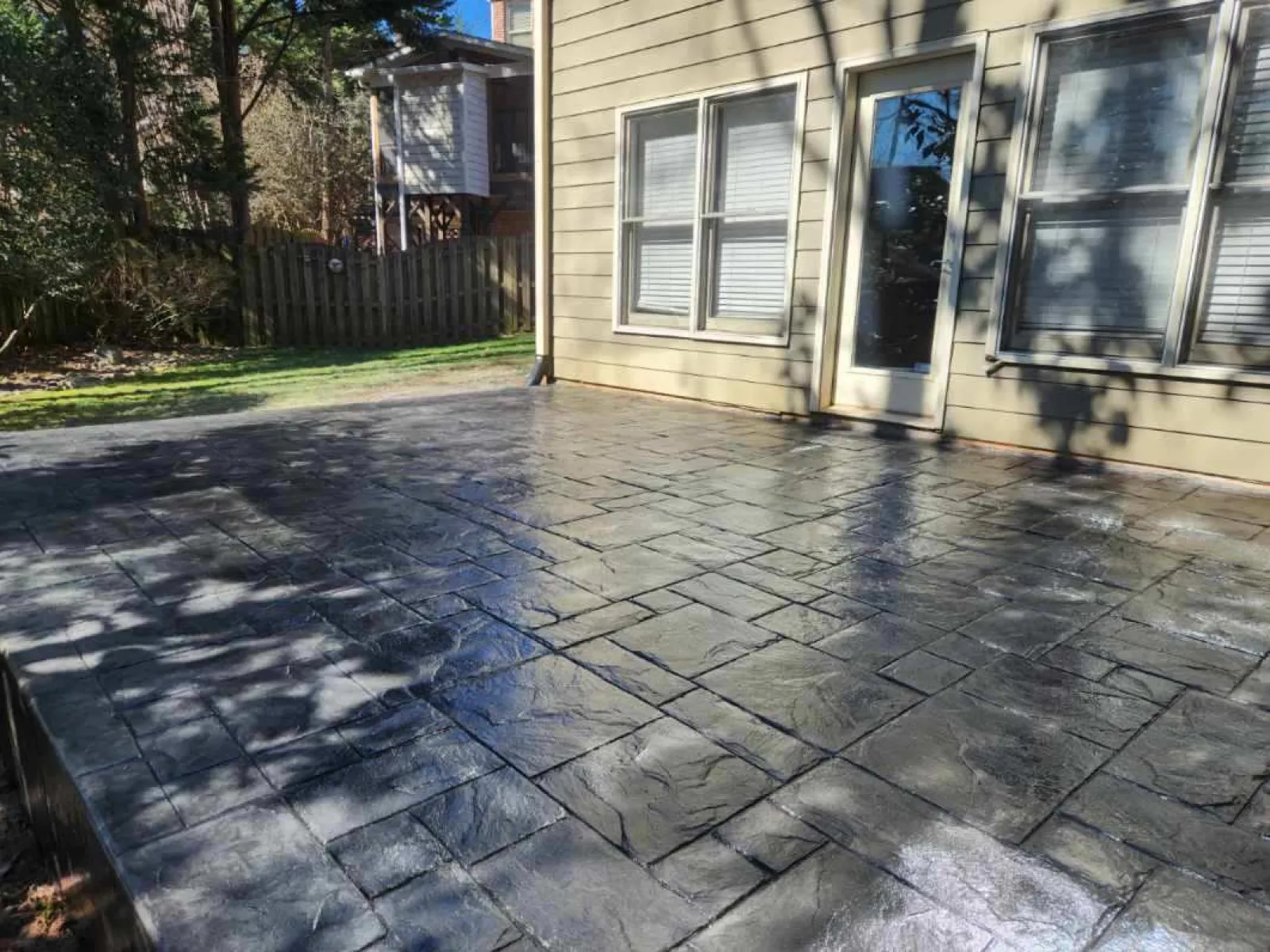 Stylish stamped concrete patio with a sleek gray finish beside a home, featuring sunlight and shadows.