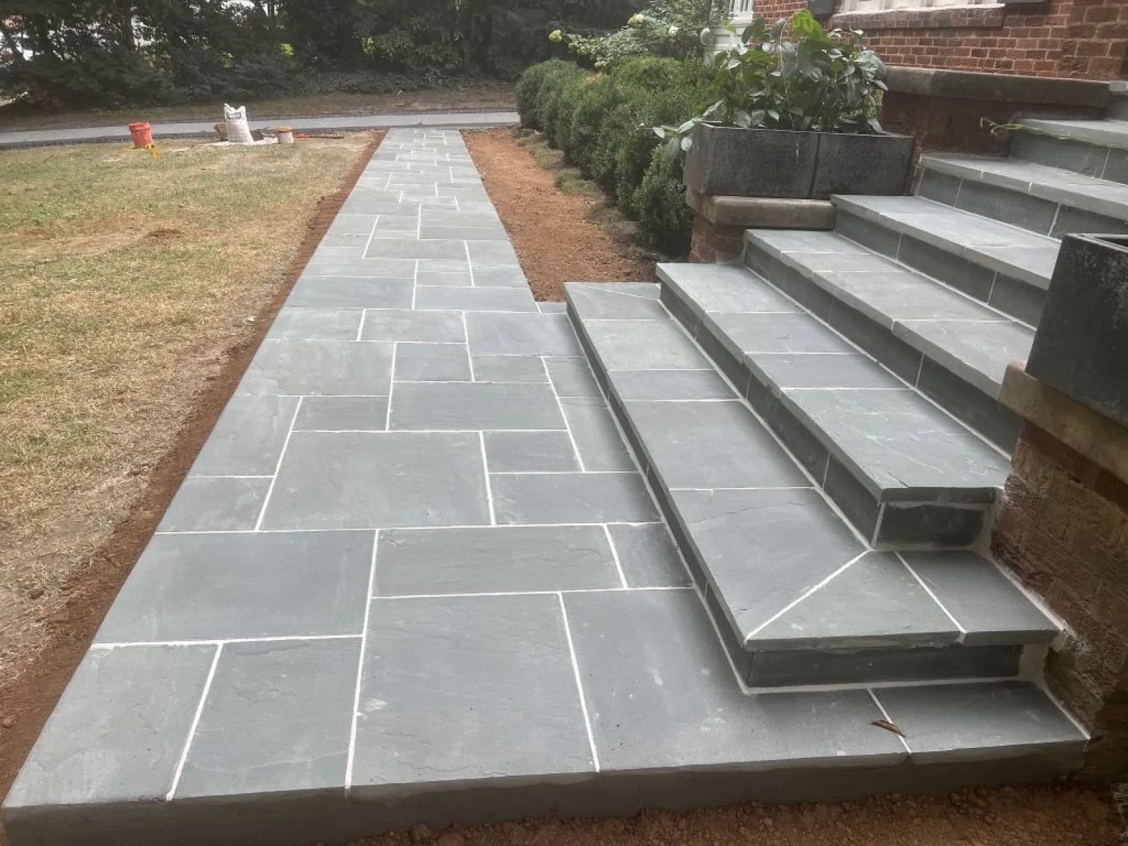 Newly installed gray stone walkway and stairs leading to a brick house with green shrubs nearby.