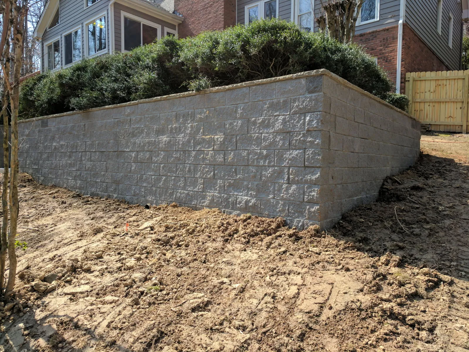 Newly constructed gray concrete retaining wall supporting bushes beside a house with exposed dirt ground.