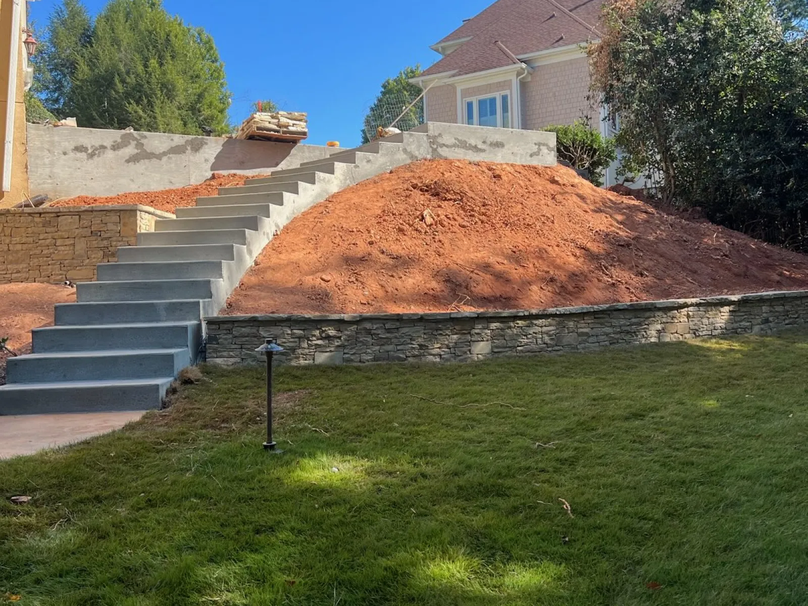 A newly constructed landscape featuring a stone retaining wall and steps leading up a sloped dirt hill under a clear blue sky.