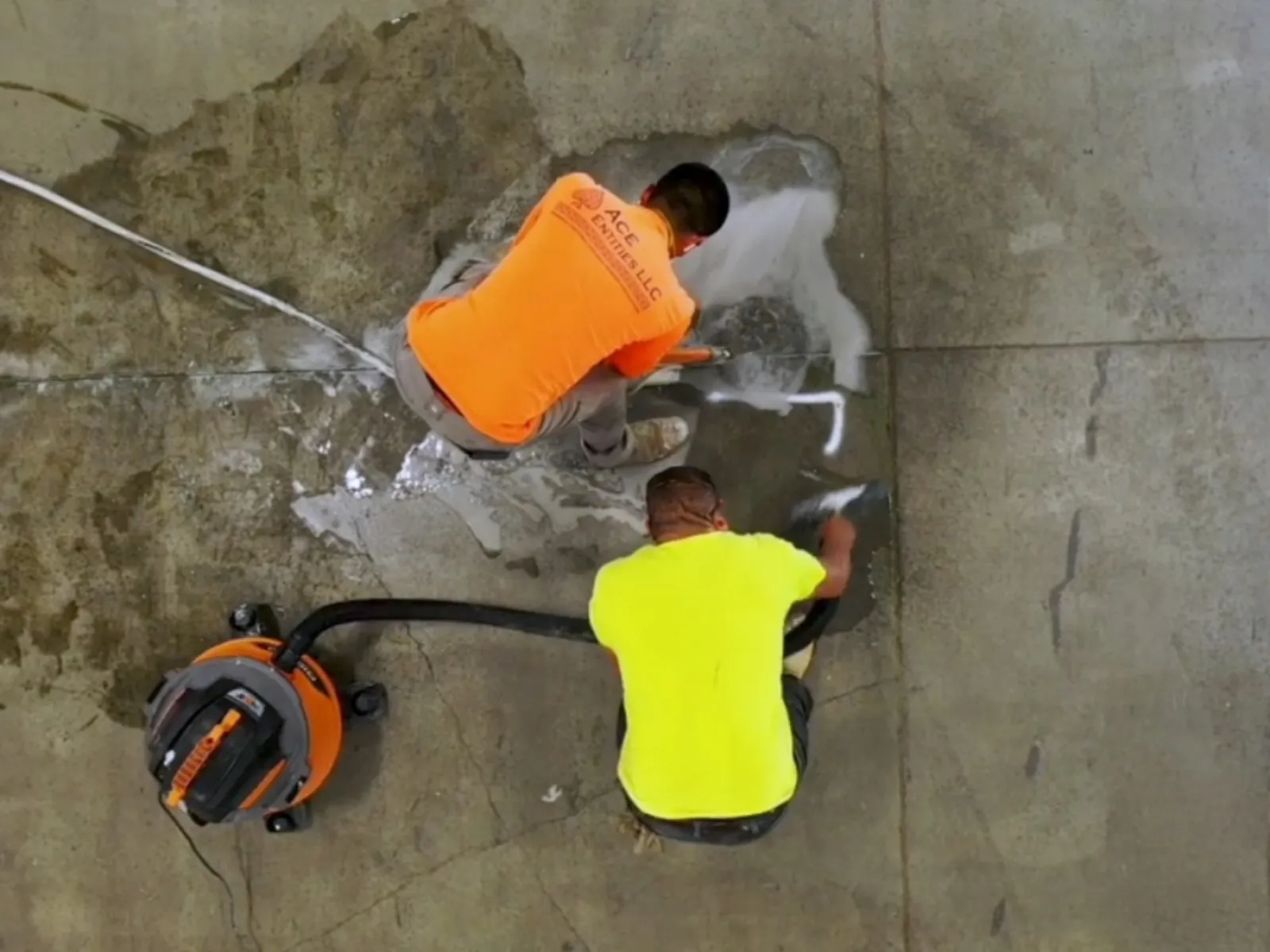 Two workers in bright shirts repairing and cleaning a cracked concrete floor using tools and vacuum equipment.