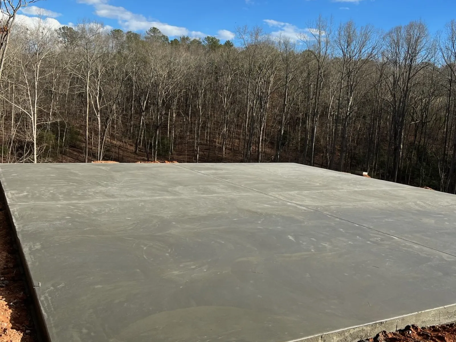 Freshly poured concrete slab foundation in a clearing surrounded by bare trees under a blue sky with clouds.