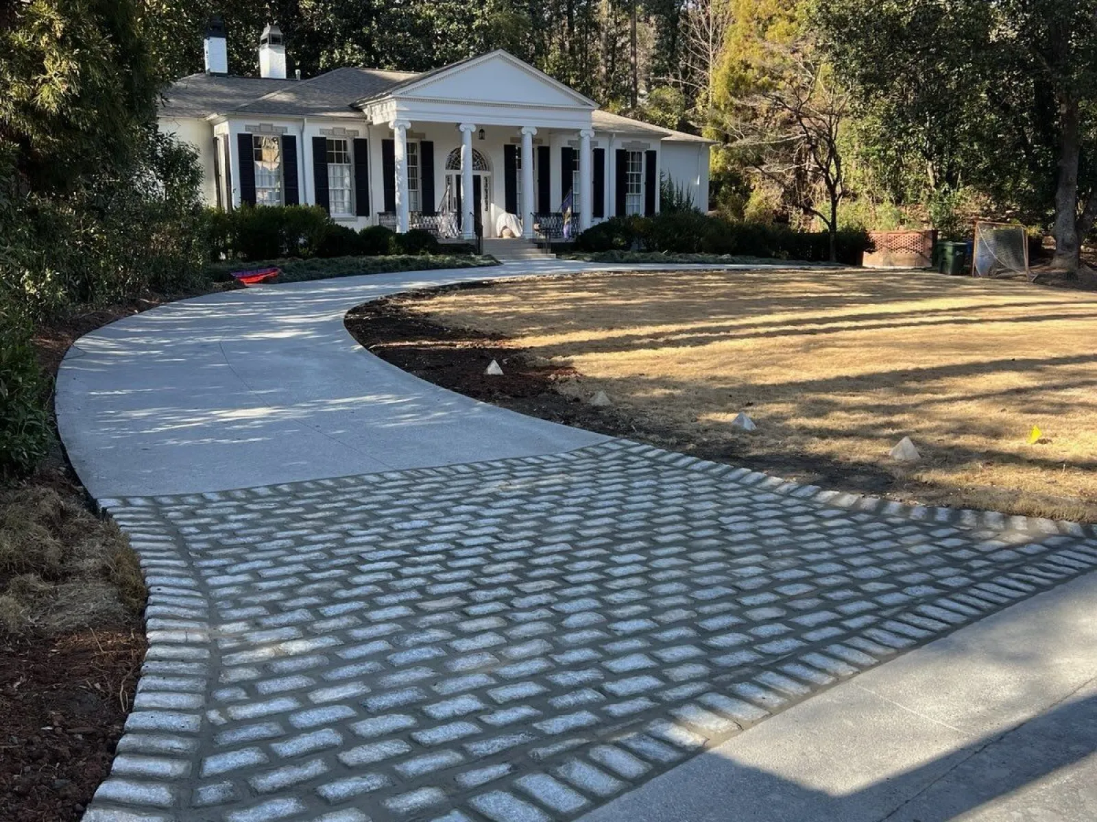 Curved driveway with cobblestone and concrete paving leading to a white colonial-style house surrounded by trees.