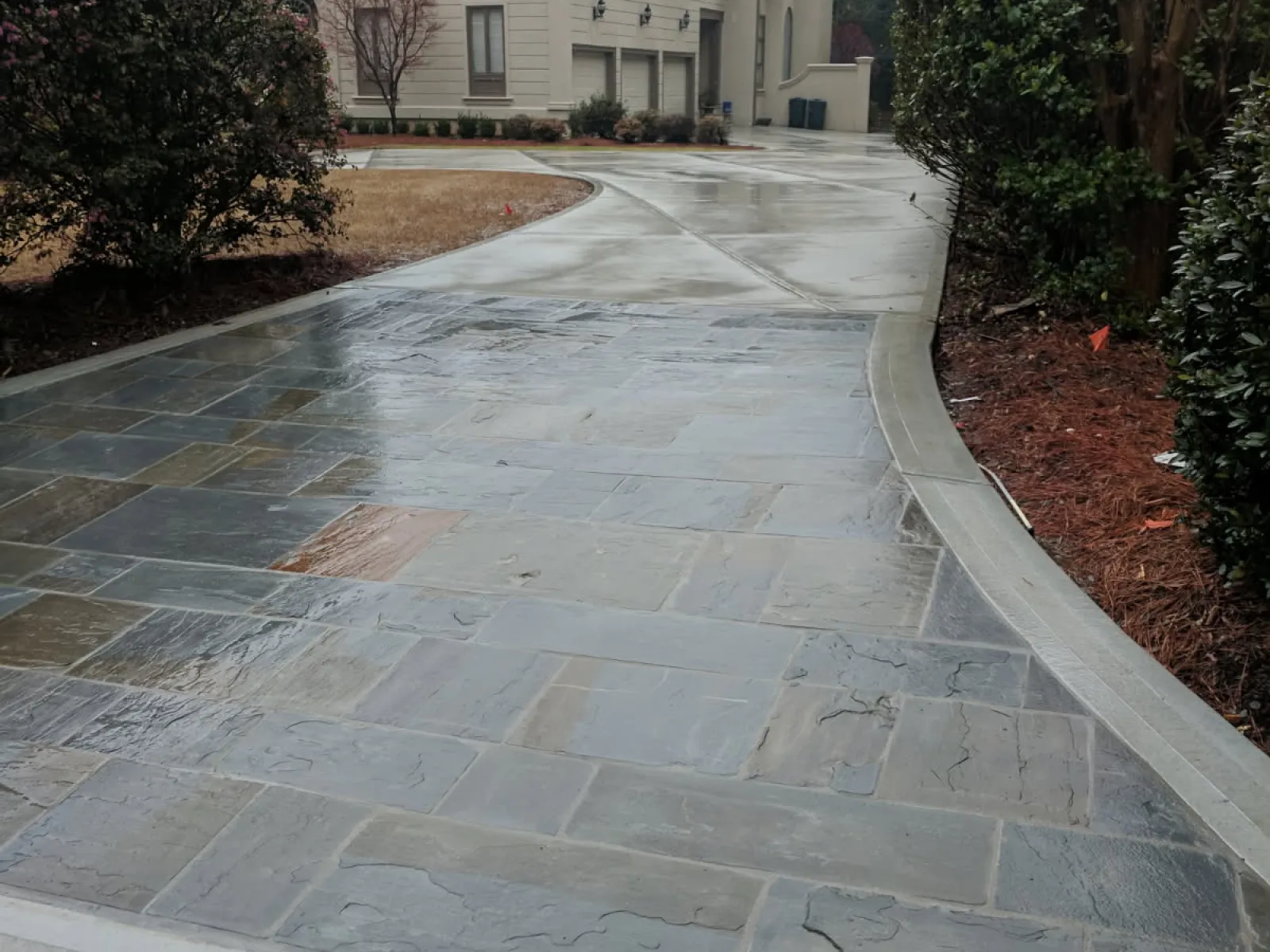 Wet stone driveway with concrete border leading to a large beige house surrounded by trees and shrubs.