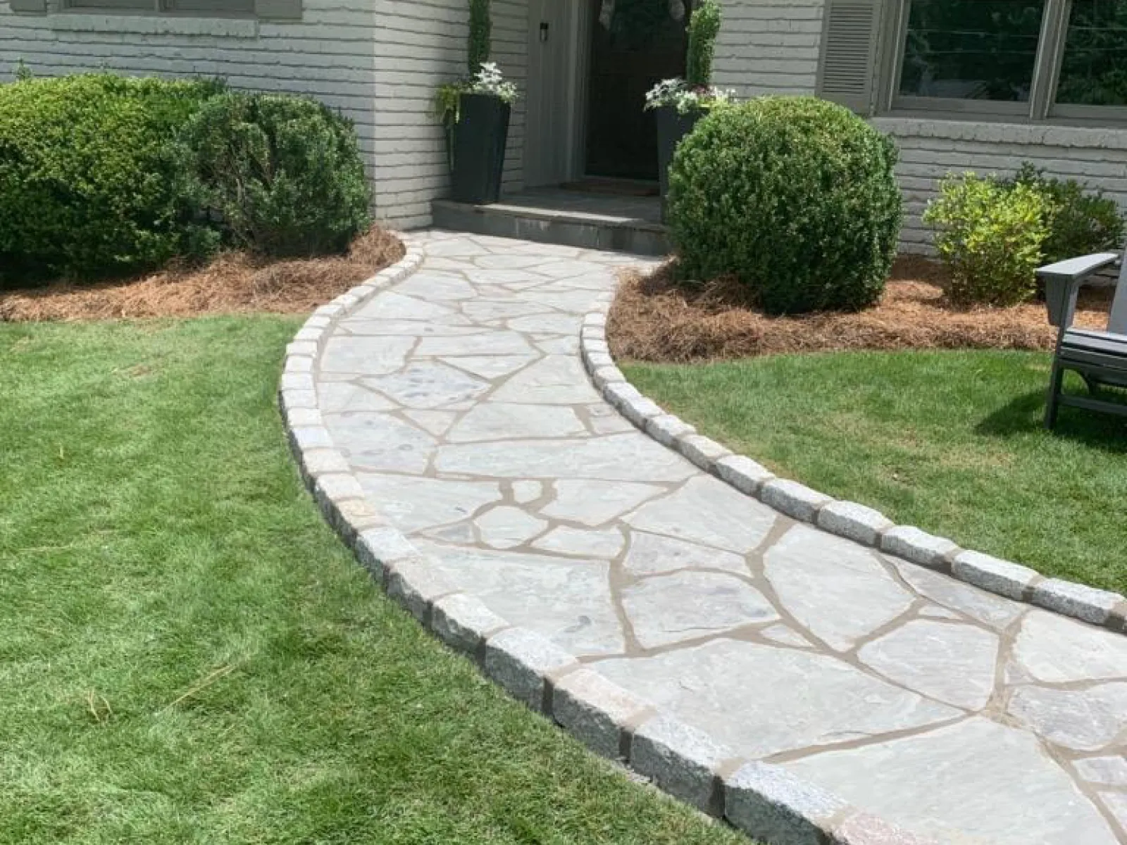 A beautifully landscaped pathway leads to a charming white brick house surrounded by green bushes.
