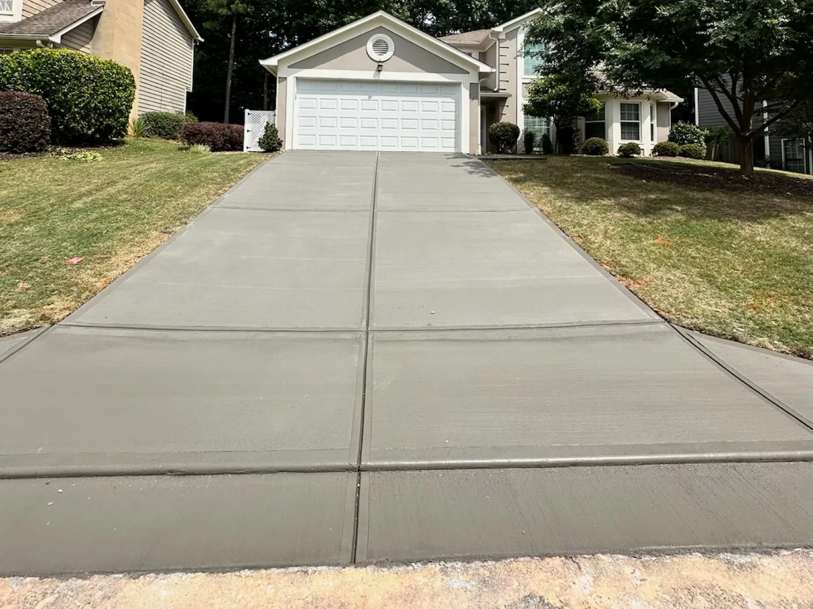 Freshly poured concrete driveway leading to a two-car garage with surrounding green lawn and trees.