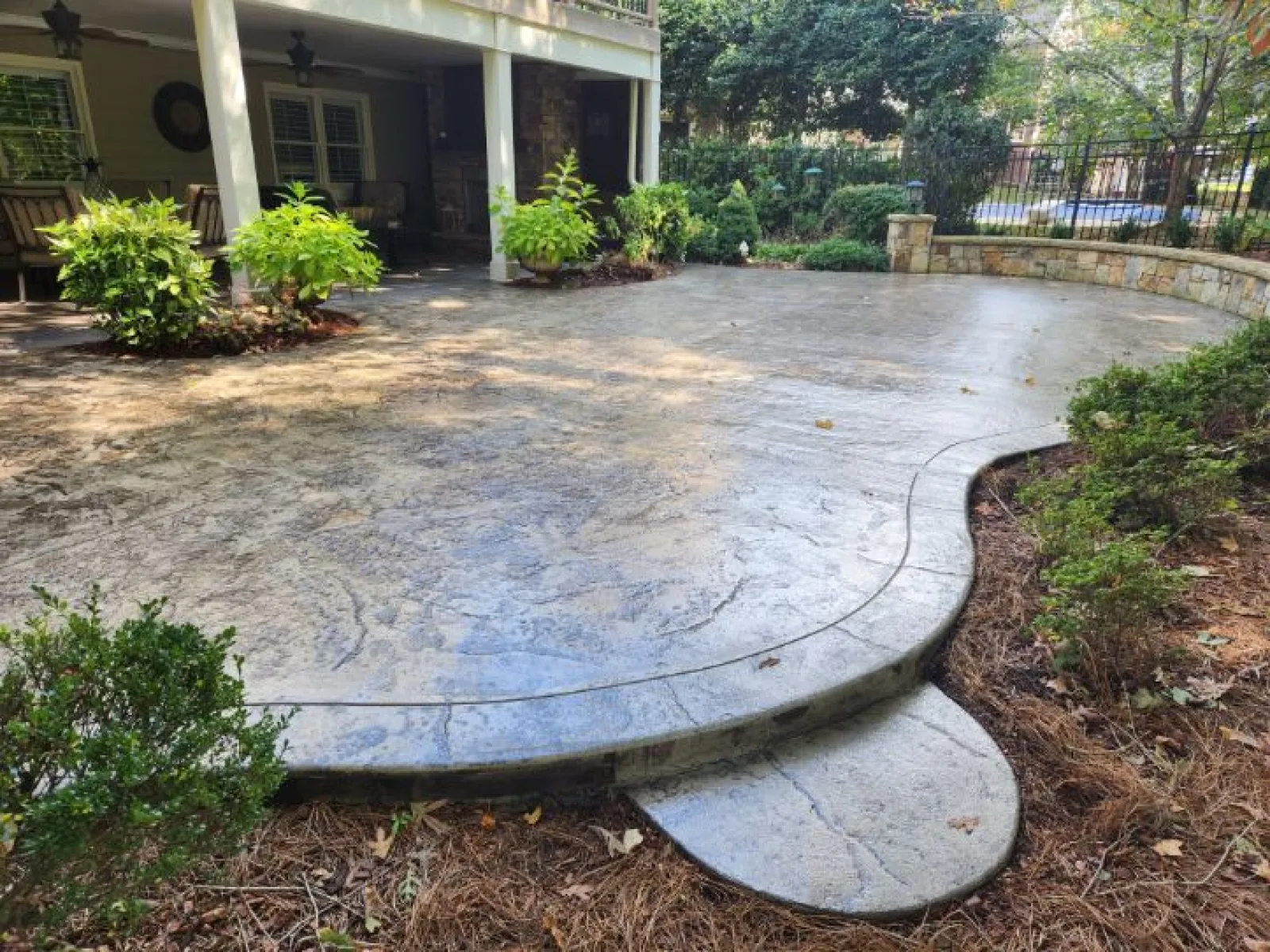 Beautiful stamped concrete patio surrounded by lush greenery and trees in a residential backyard setting.
