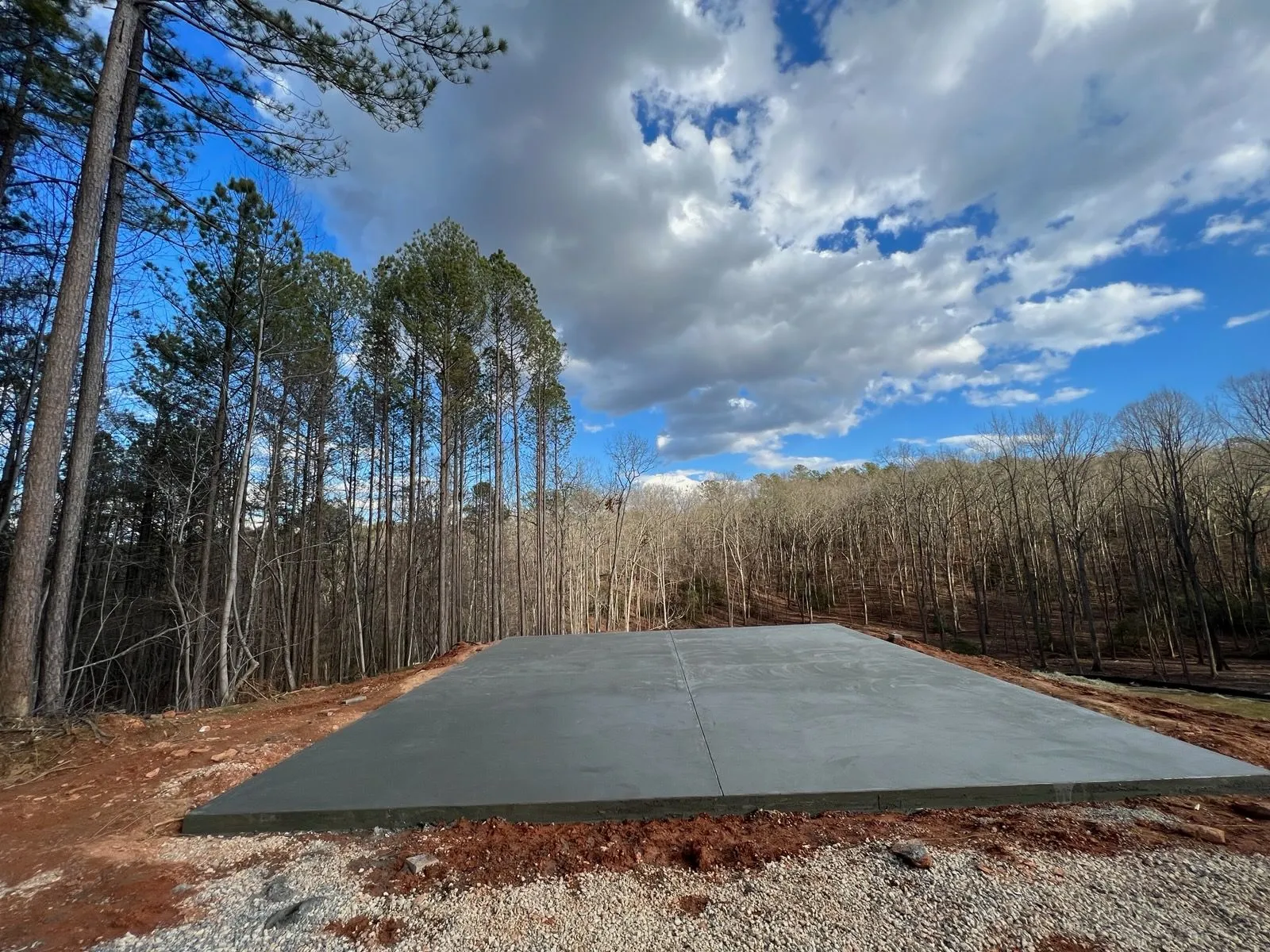 New concrete foundation slab in clearing surrounded by bare trees and a cloudy blue sky.