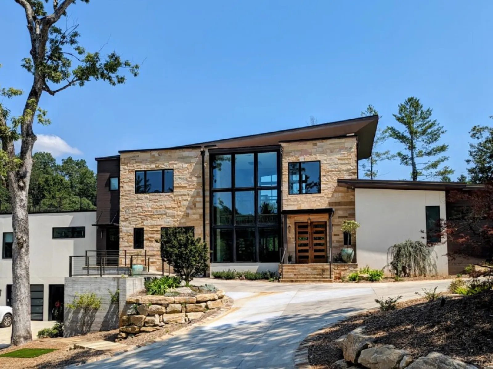 Modern luxury home with large windows, natural stone facade, and landscaped driveway surrounded by trees.