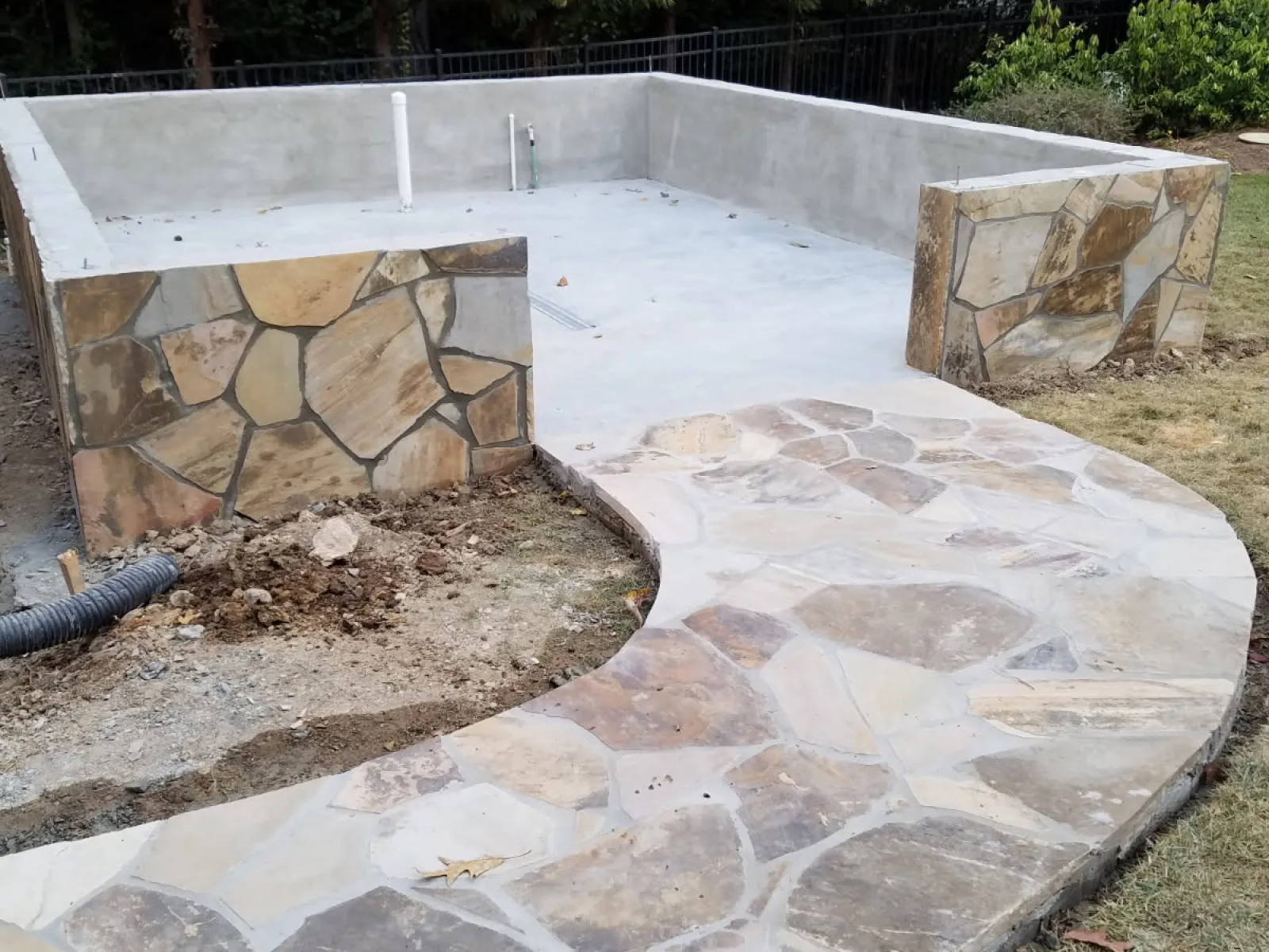 Curved stone pathway leading to a partially constructed concrete pool with stone-faced walls in a backyard.