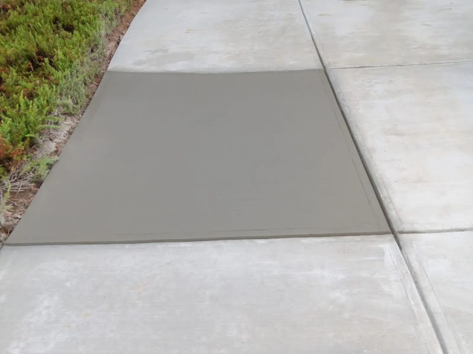 Freshly poured gray concrete on a driveway, with surrounding greenery and textured concrete patterns.