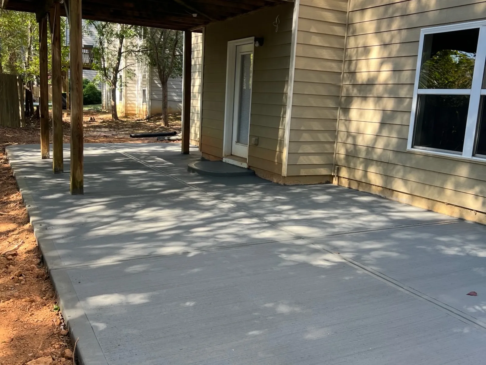 Freshly poured gray concrete patio under a deck, surrounded by trees and a well-maintained yard.