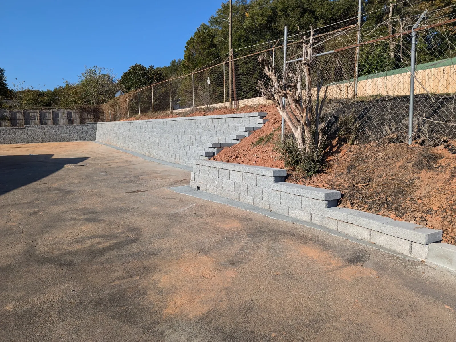 Concrete retaining walls with steps lining a dirt slope beside an asphalt area under clear blue sky.