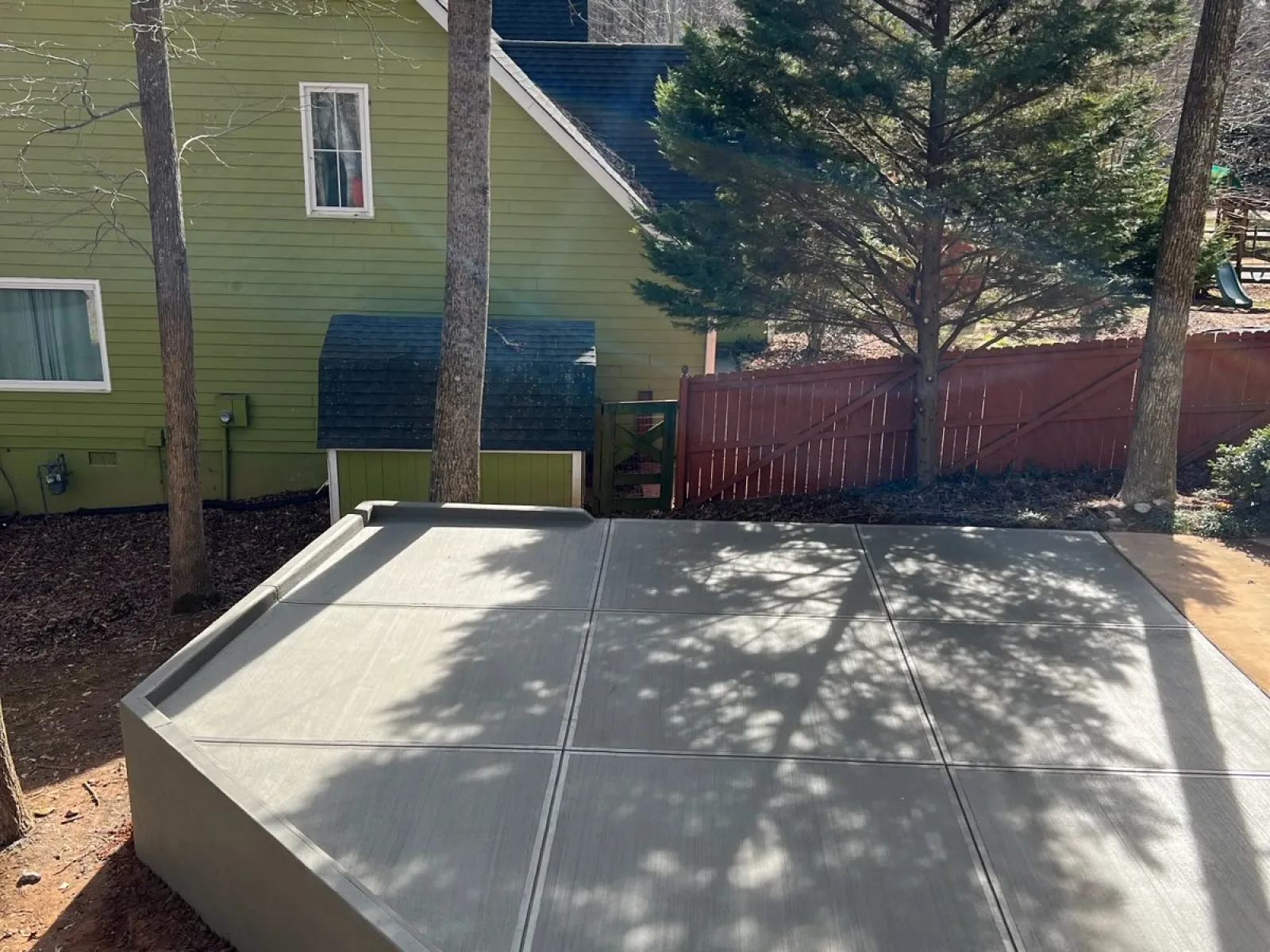Newly poured concrete slab patio in backyard with trees, green house, and red wooden fence in background.