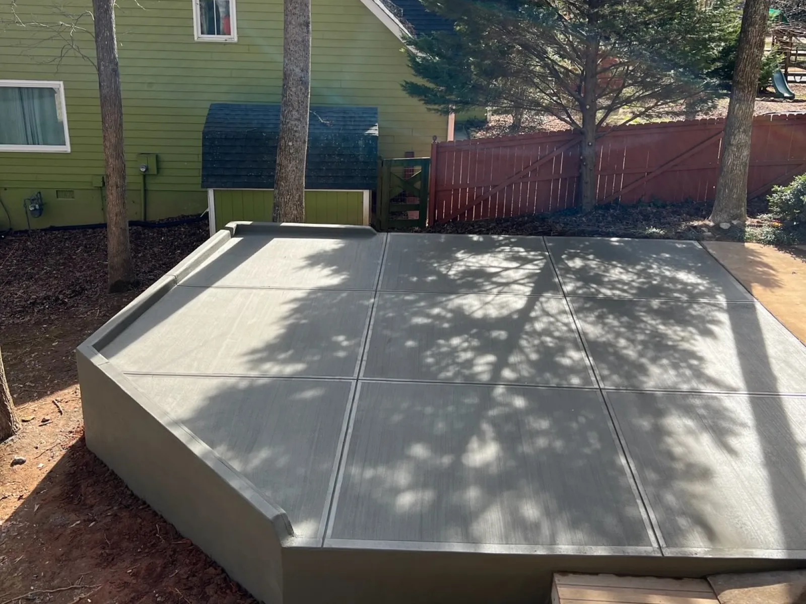 Newly poured concrete slab patio in backyard with trees, green house, and red wooden fence in background.