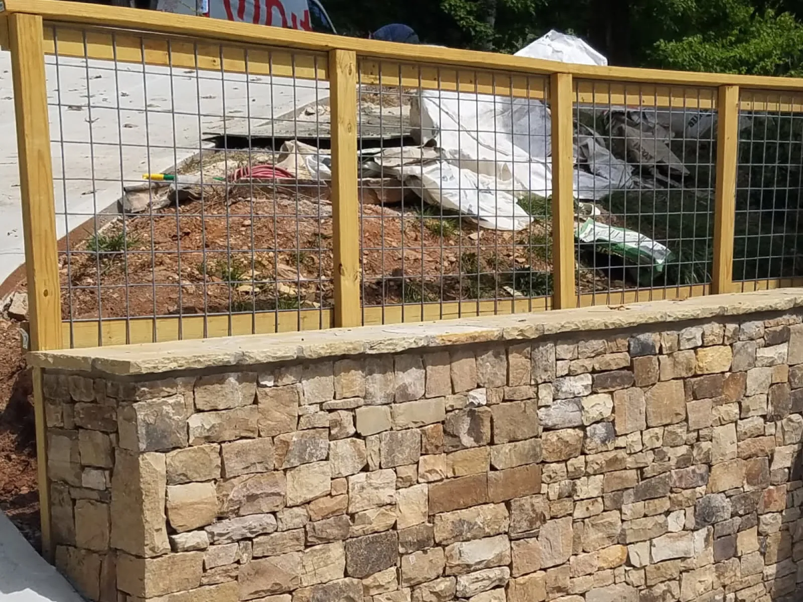 Stone retaining wall with a wood and wire mesh fence above on a concrete driveway corner under sunlight.