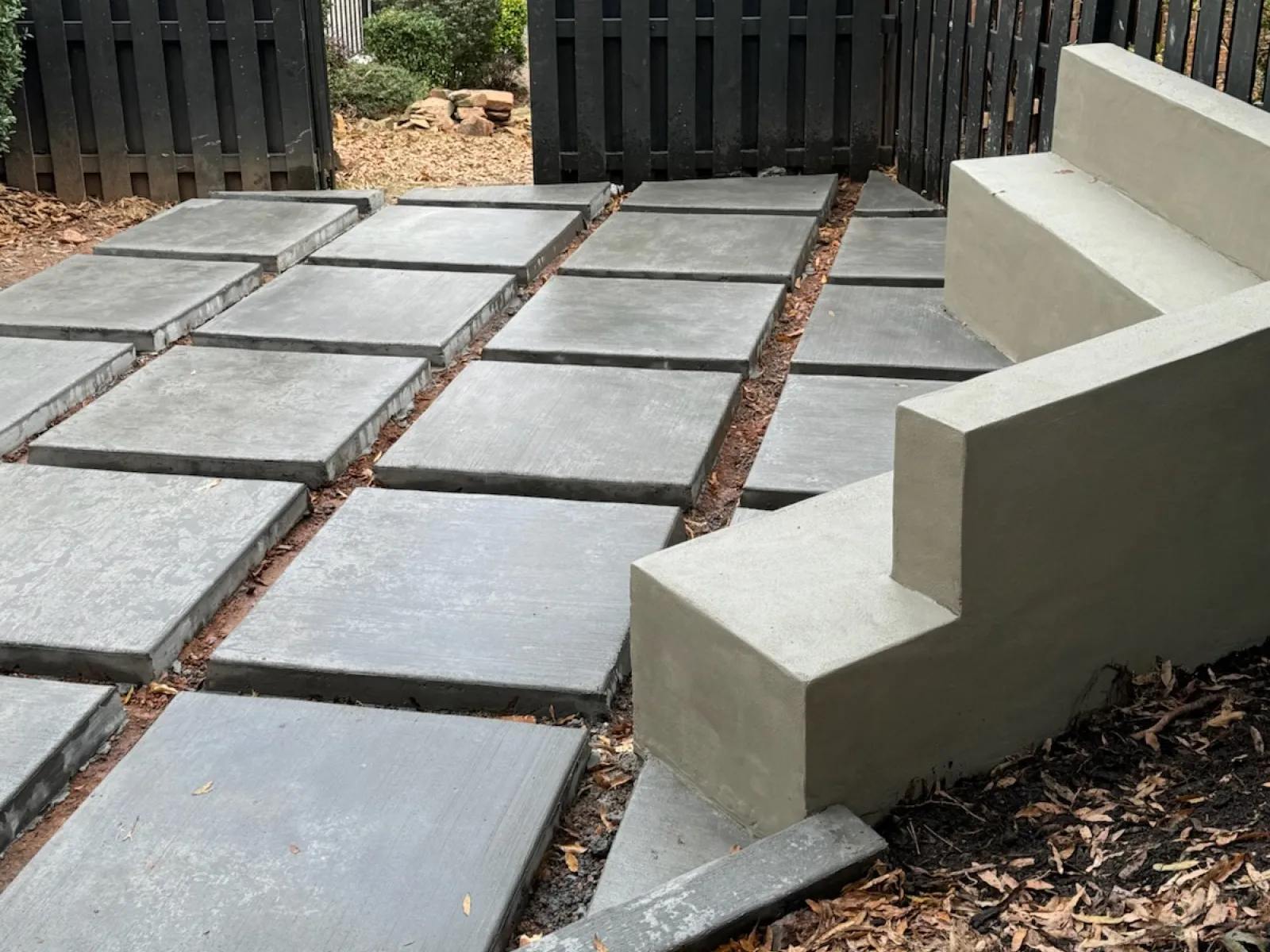 Outdoor space with large square concrete pavers and built-in concrete seating against a black fence.