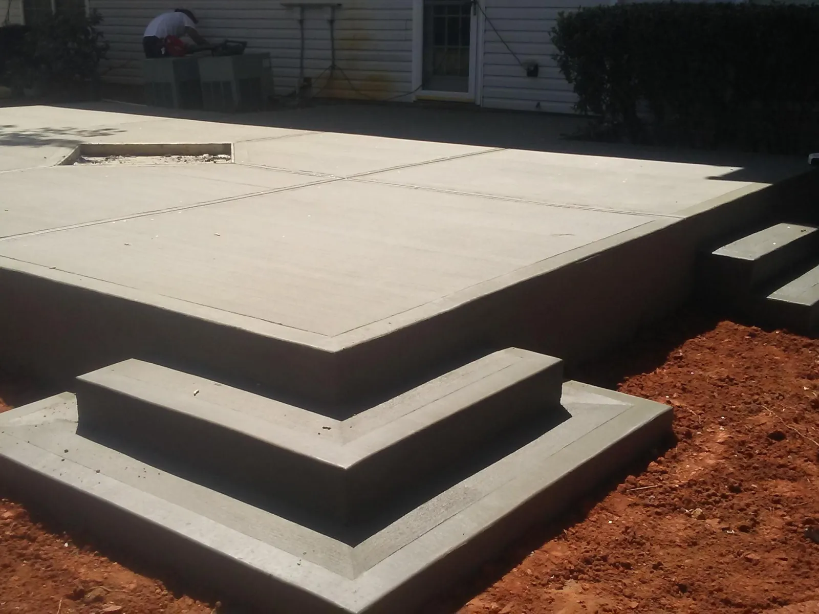 Newly poured concrete patio with multi-level steps in a backyard garden area beside a house.