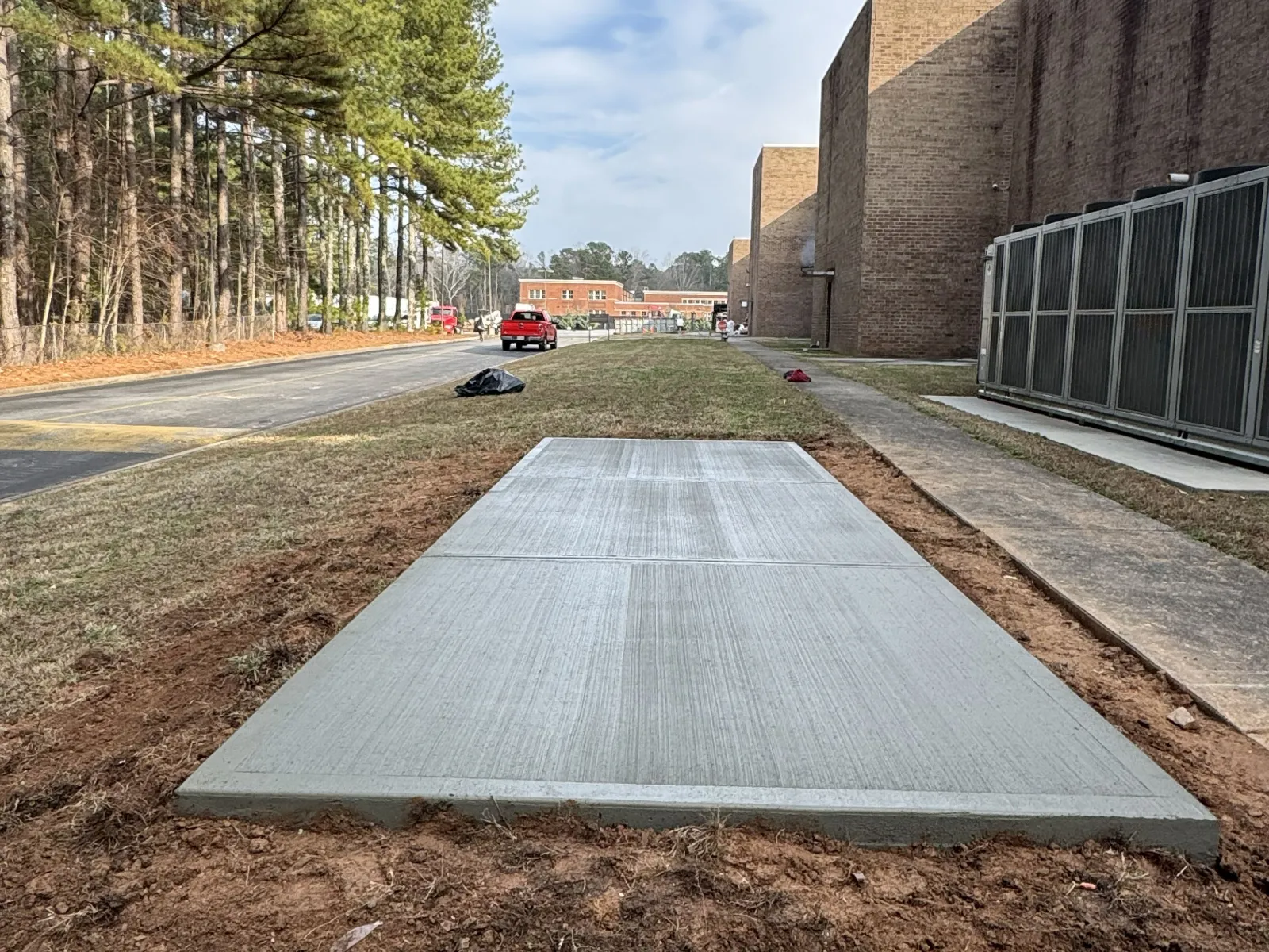 Newly poured concrete slab next to a brick building with trees and road in background under cloudy sky.