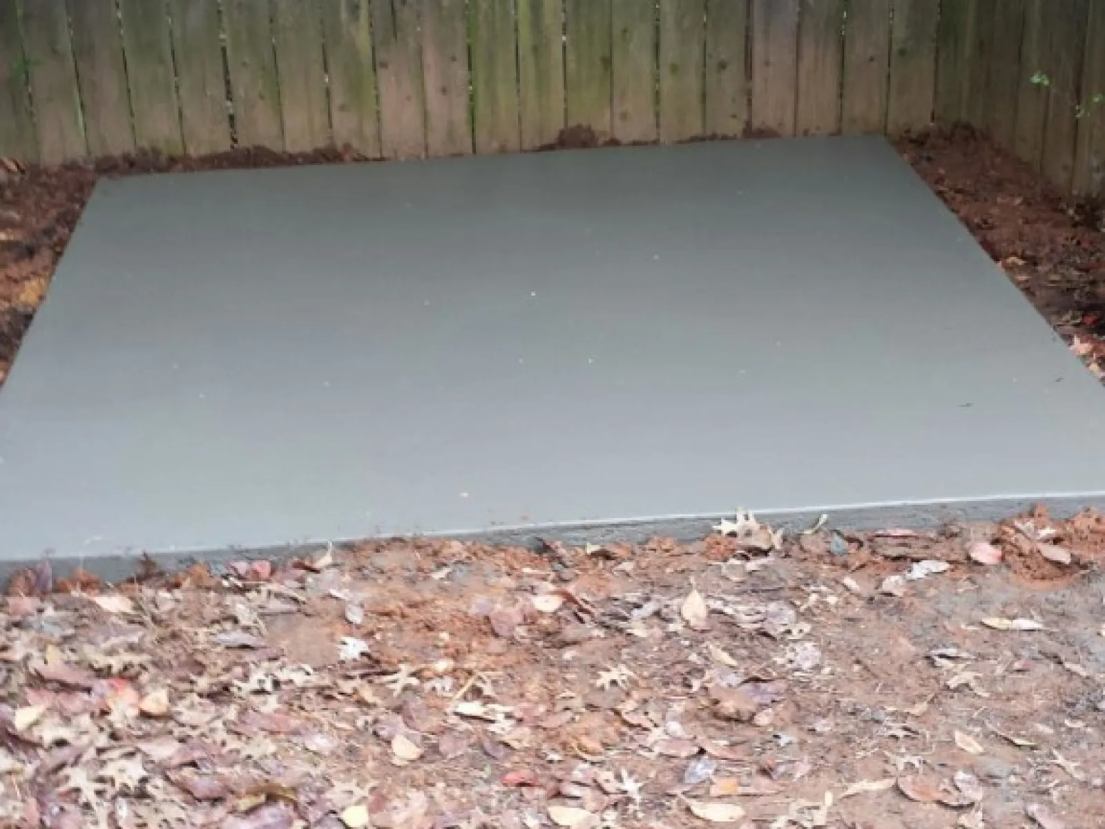 Gray concrete slab installed in a backyard, surrounded by fallen leaves and wooden fence.