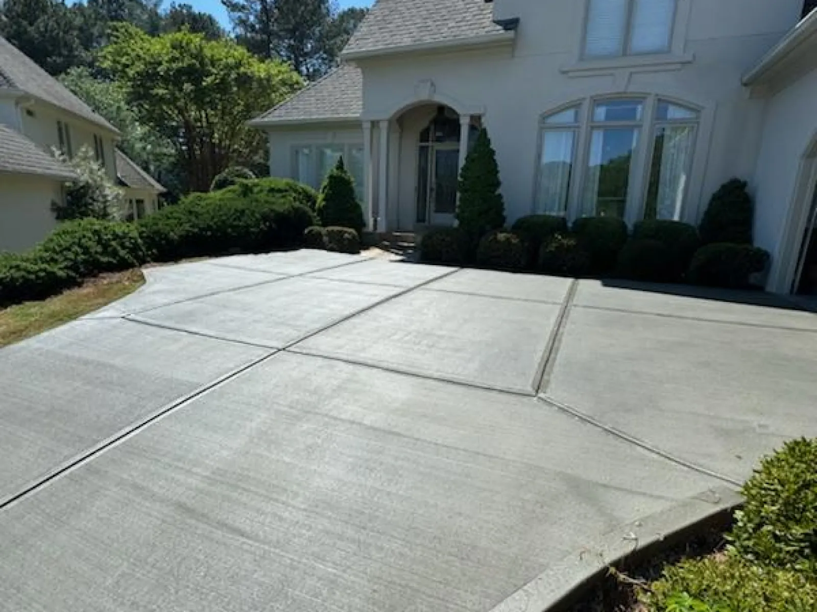 Freshly poured concrete driveway leading to a large white residential house with manicured shrubs and clear blue sky.