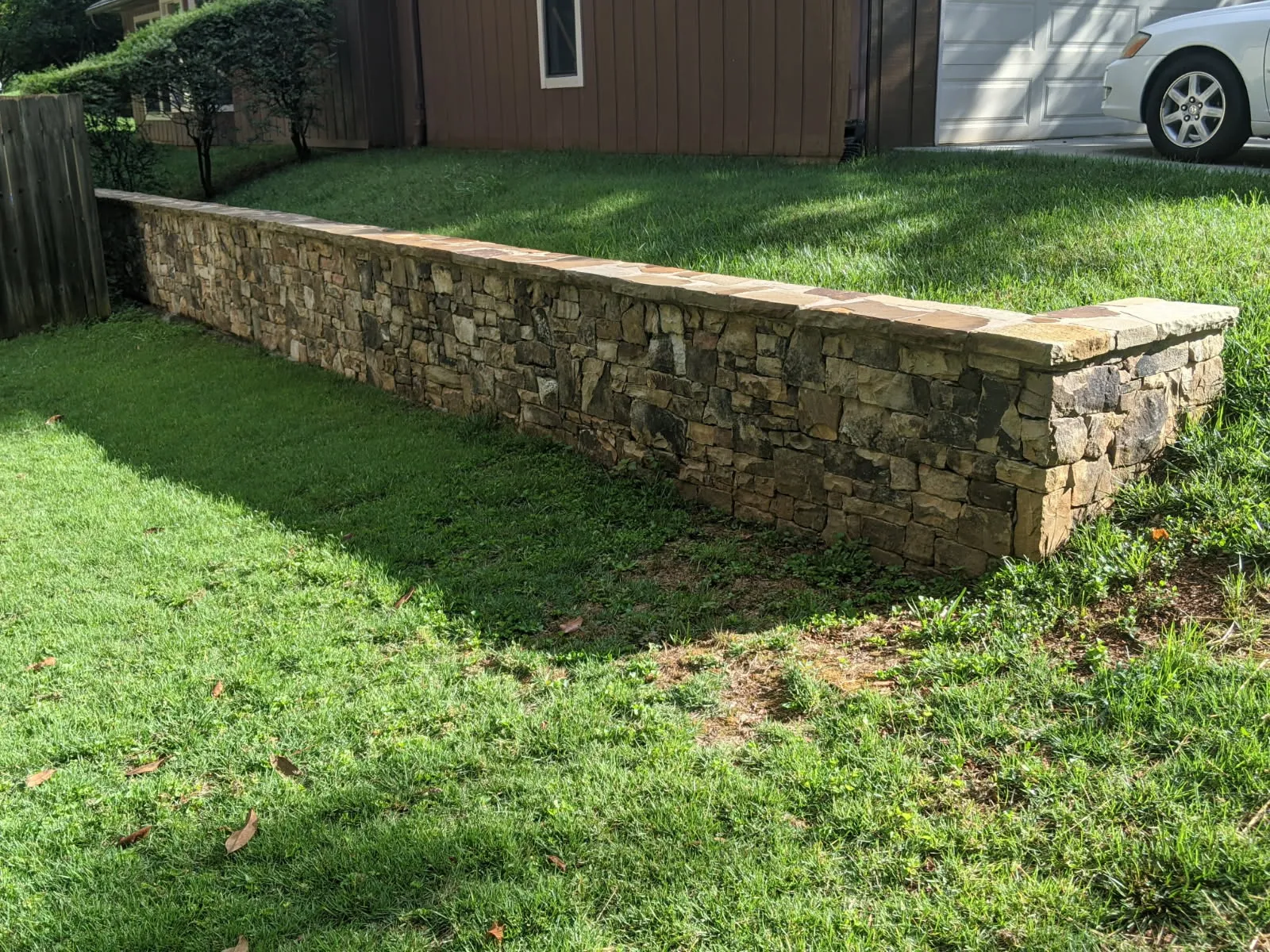 Long stone retaining wall separates two grassy levels in a residential yard with driveway and car in background
