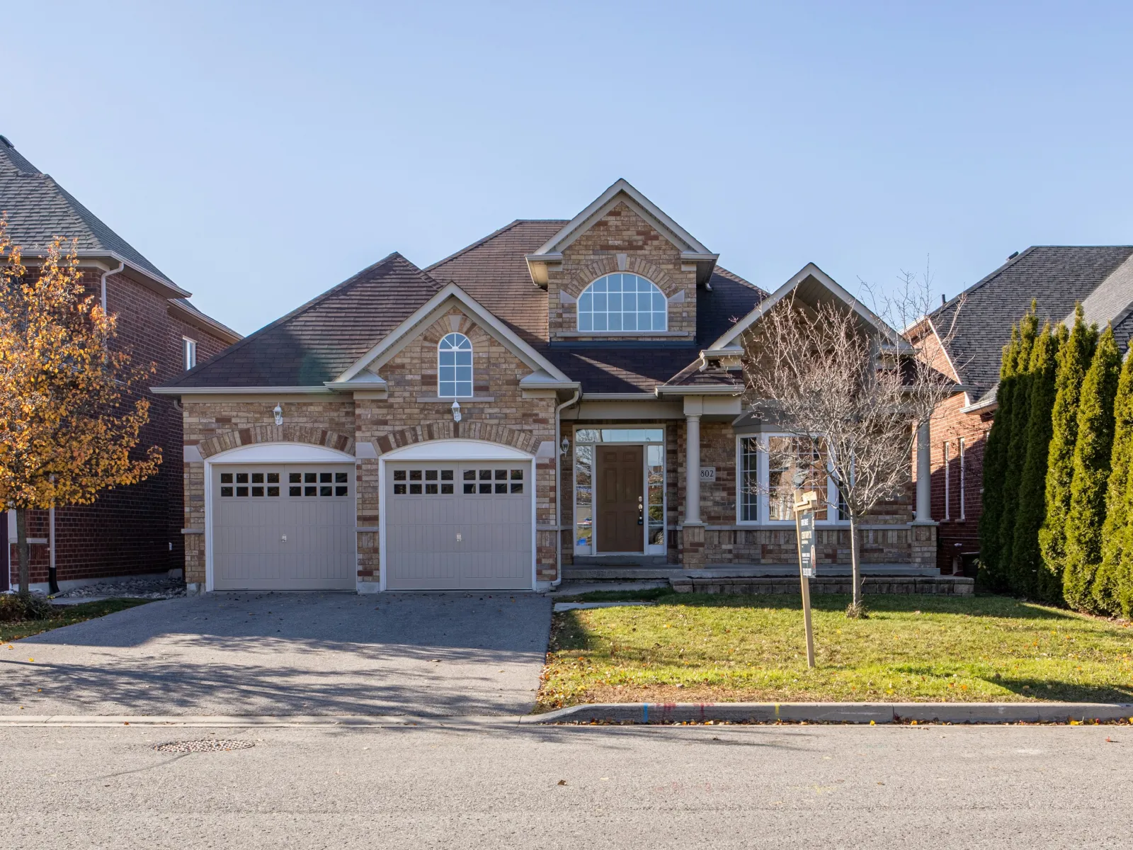Brick suburban house with double garage, arched windows, green lawn, and trees under clear blue sky.