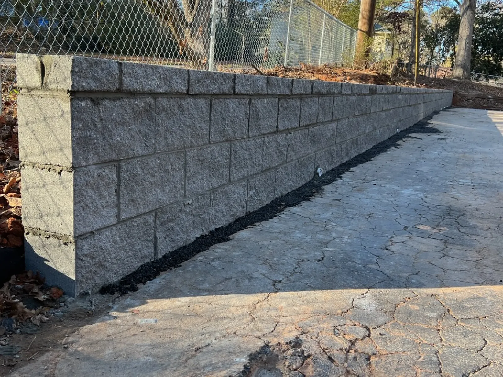 Concrete retaining wall next to cracked asphalt driveway under clear blue sky in a wooded area.