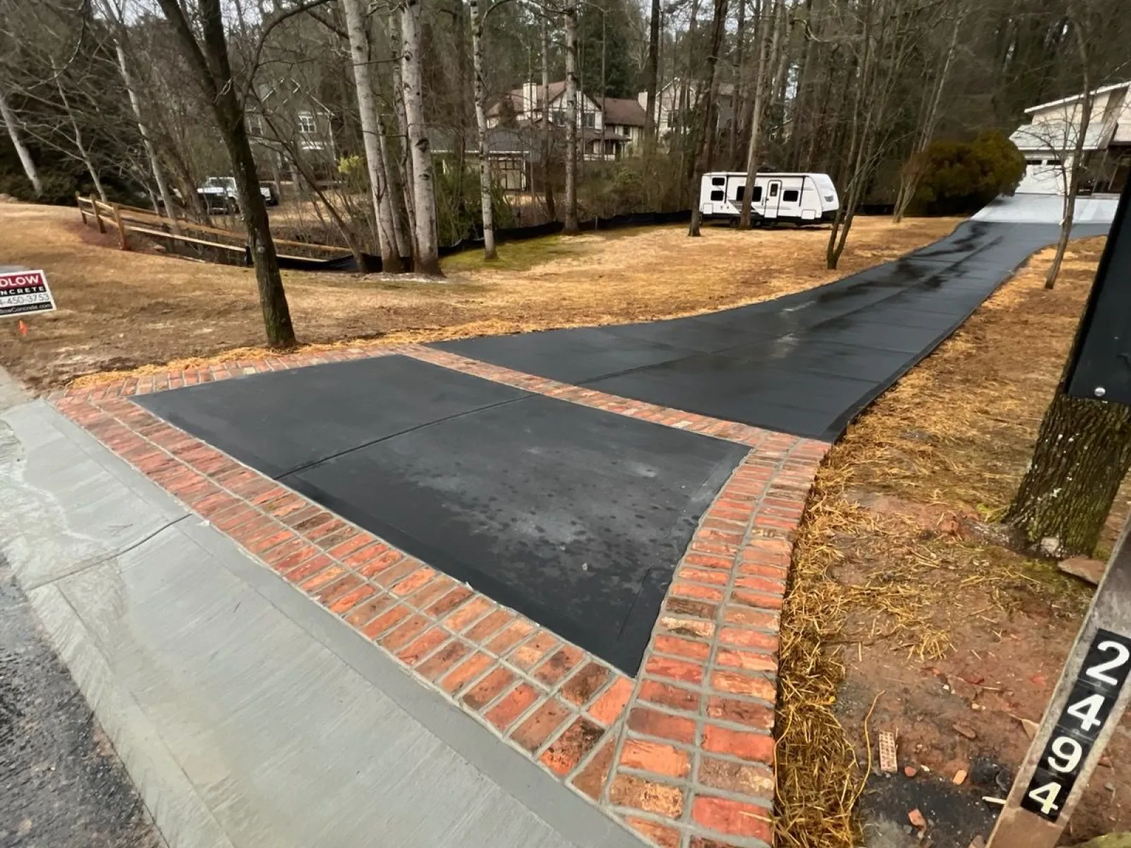 Newly paved black driveway with red brick border and concrete ramp leading to a house in a wooded area.