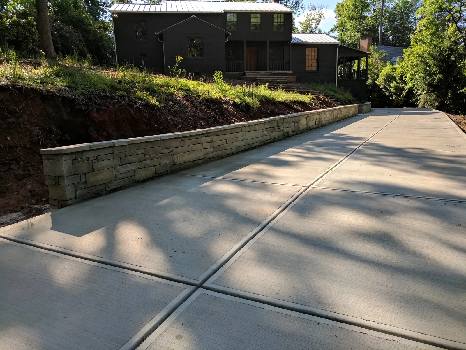 Wide concrete driveway with stone retaining wall leading to a modern dark house surrounded by trees