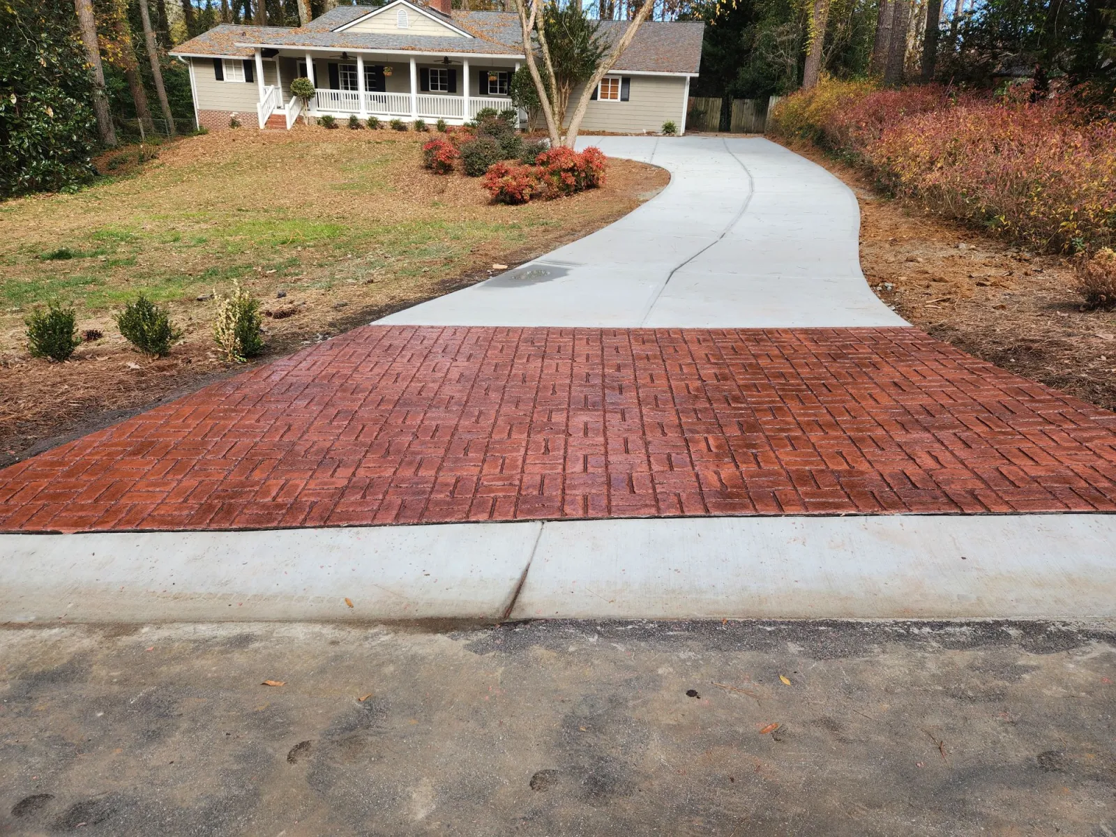 Curved concrete driveway with red brick-patterned section leading to a house surrounded by trees and landscaping.
