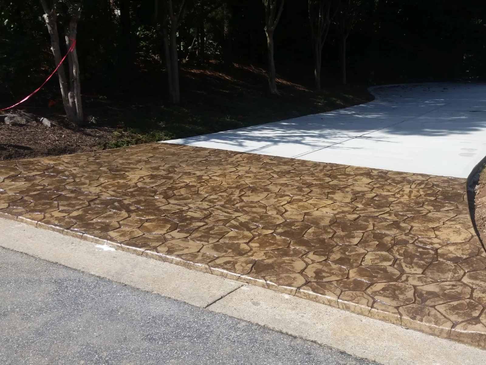 Residential driveway with stamped concrete in stone pattern transitioning to smooth white concrete surrounded by trees.
