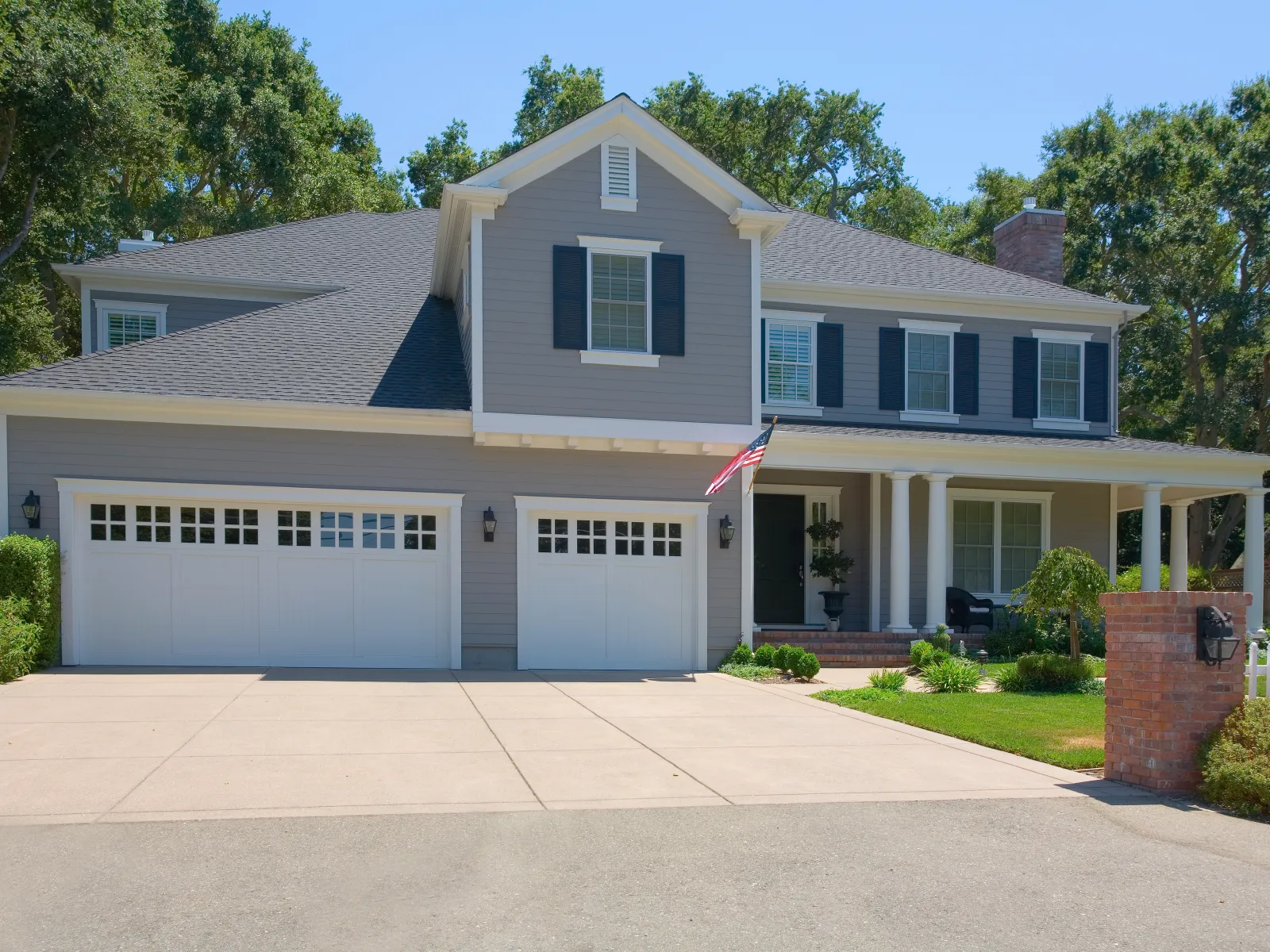 Beautiful gray suburban house with two garage doors and American flag, surrounded by lush greenery.