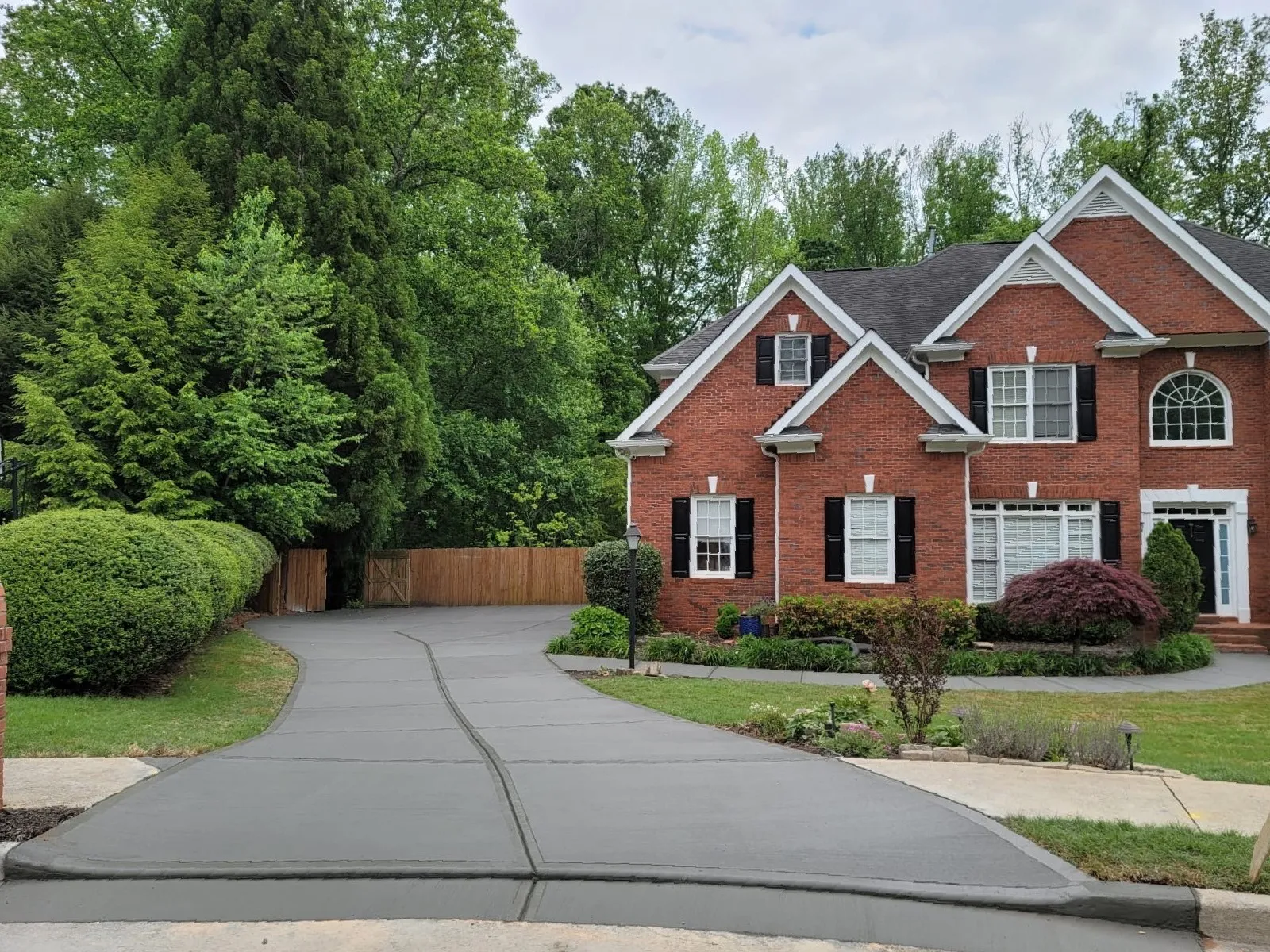 Newly paved gray concrete driveway leading to a red brick two-story house with black shutters and surrounding greenery.