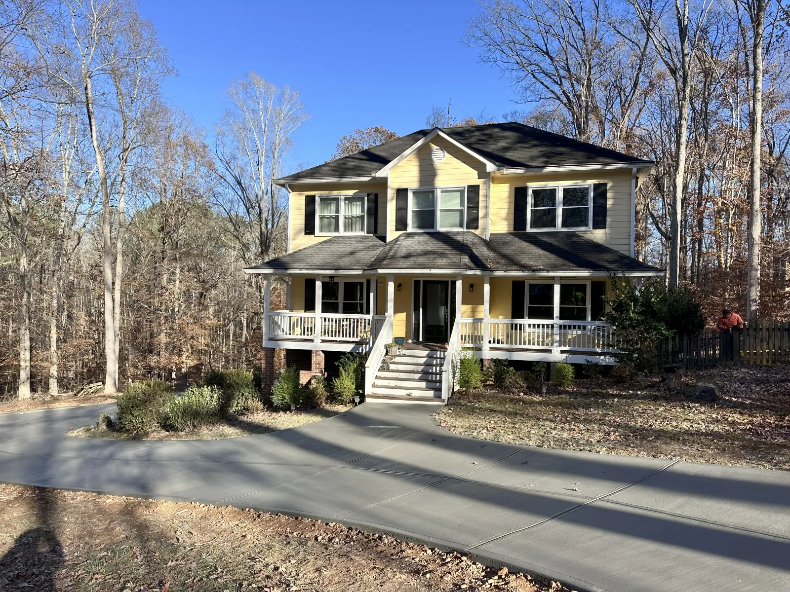 Two-story yellow house with white porch, surrounded by leafless trees and a curved concrete driveway.
