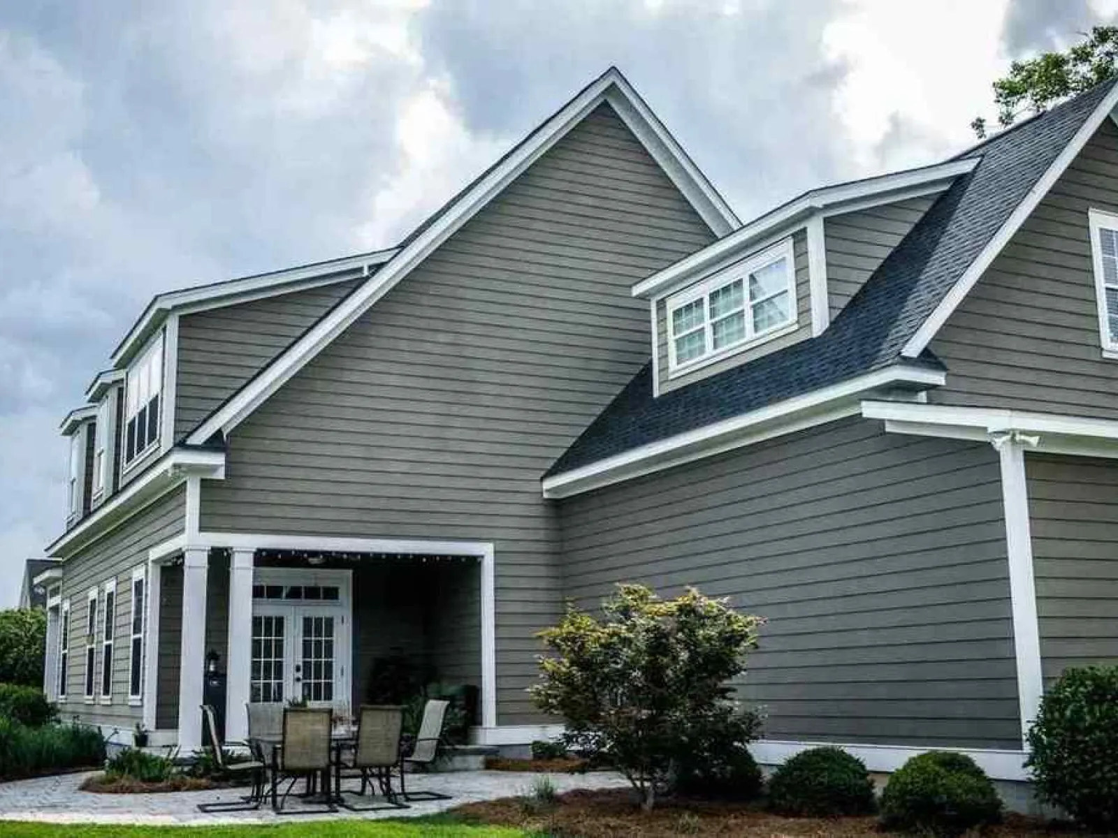 Modern gray two-story house with white trim, outdoor patio furniture, and a cloudy sky backdrop.