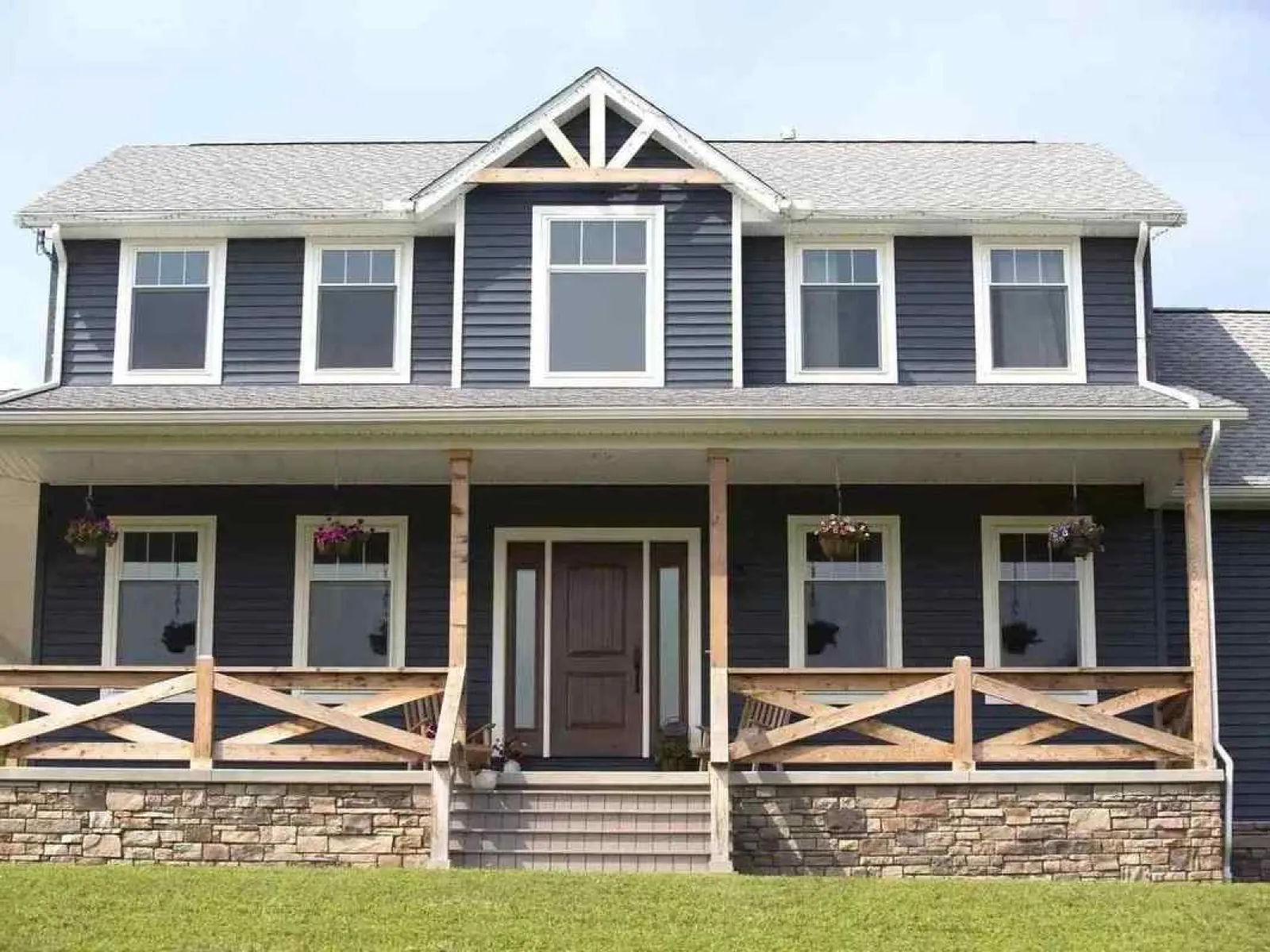 Dark blue two-story house with stone foundation, large porch, wooden railings, and white framed windows under a grey roof.
