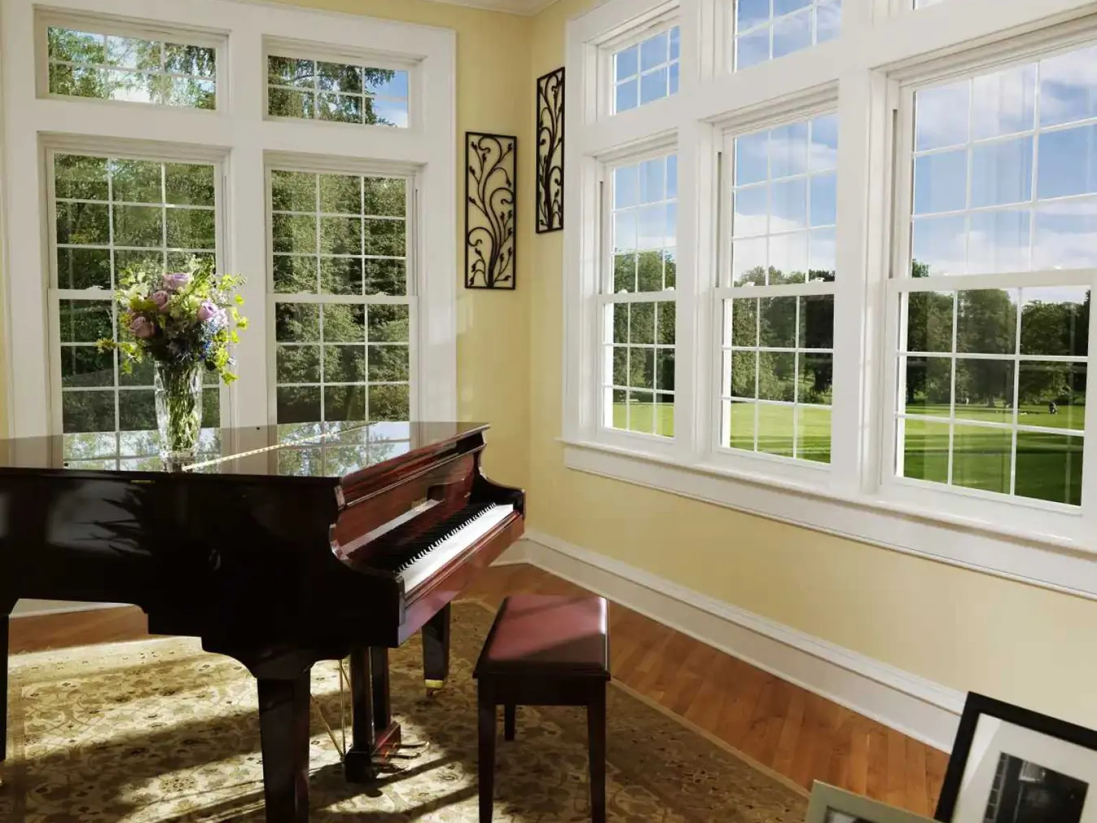 Bright room with grand piano, large windows, floral arrangement, photo frames, and patterned rug on hardwood floor