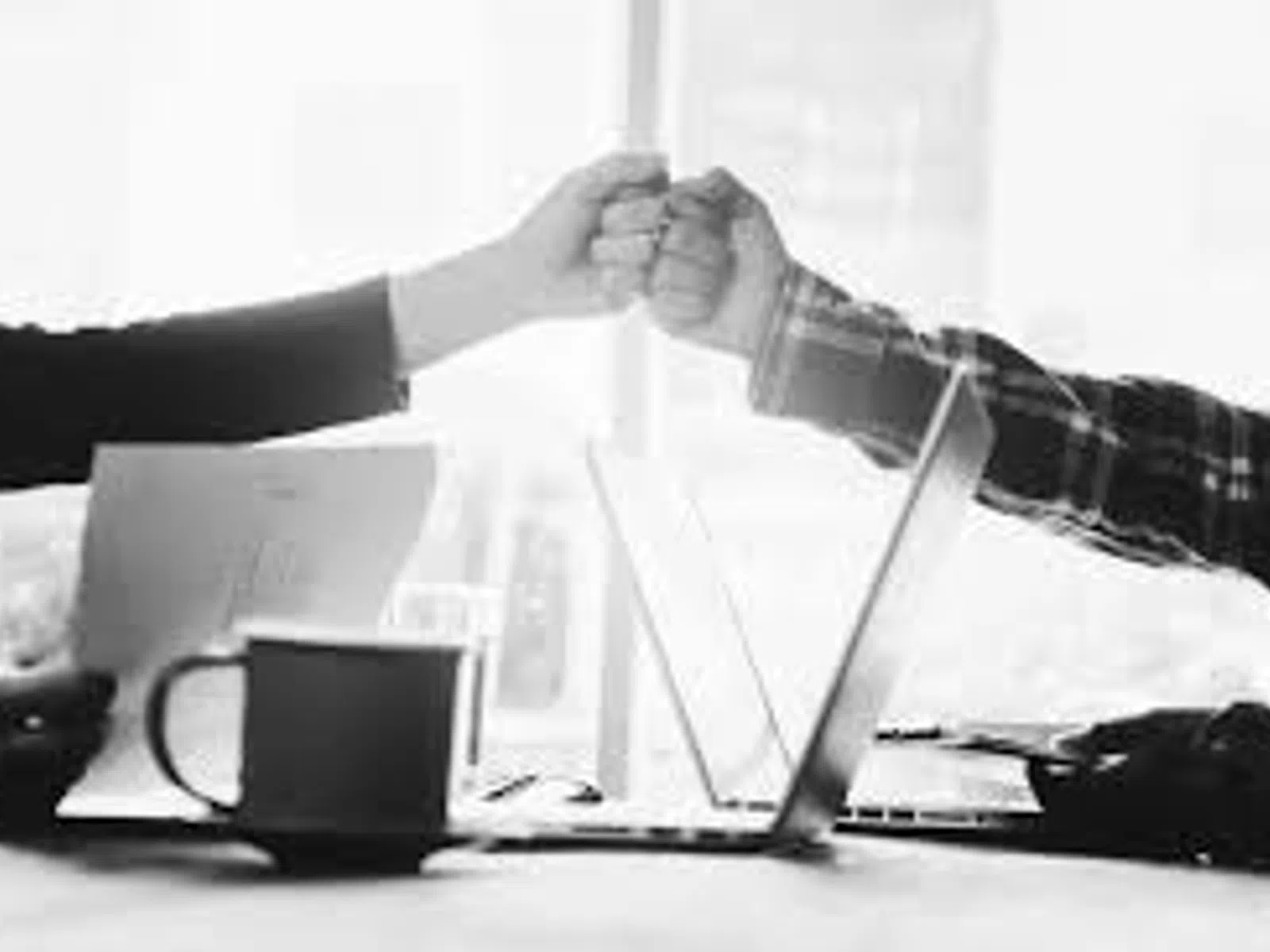 Colleagues fist bump over desk with laptop, coffee cup, and documents in a bright office setting