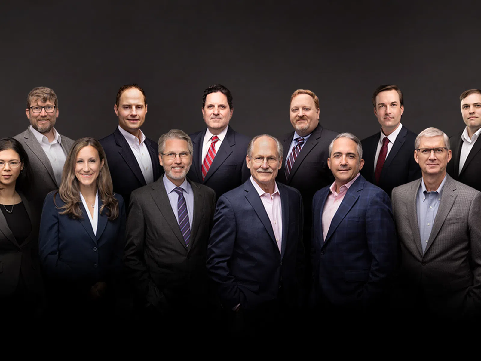 Group portrait of 15 professional men and women dressed in business attire against a dark background