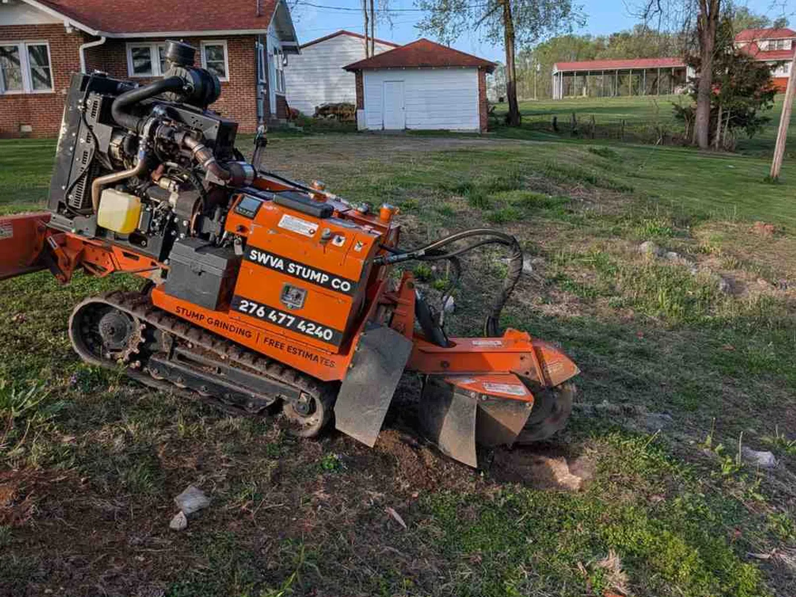 Orange stump grinding machine from SWVA Stump Co working on a residential lawn with houses and trees in background.