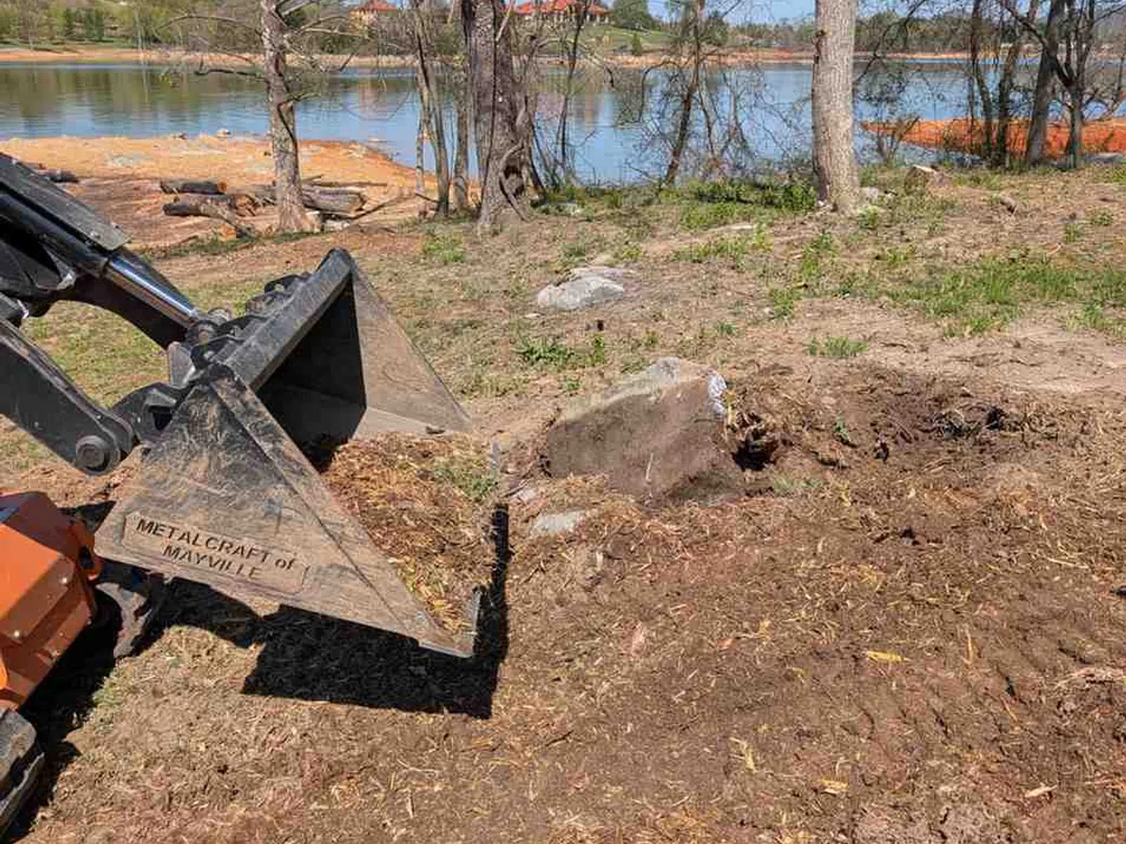 Excavator bucket clearing dirt near a lake with bare trees and a clear blue sky in the background