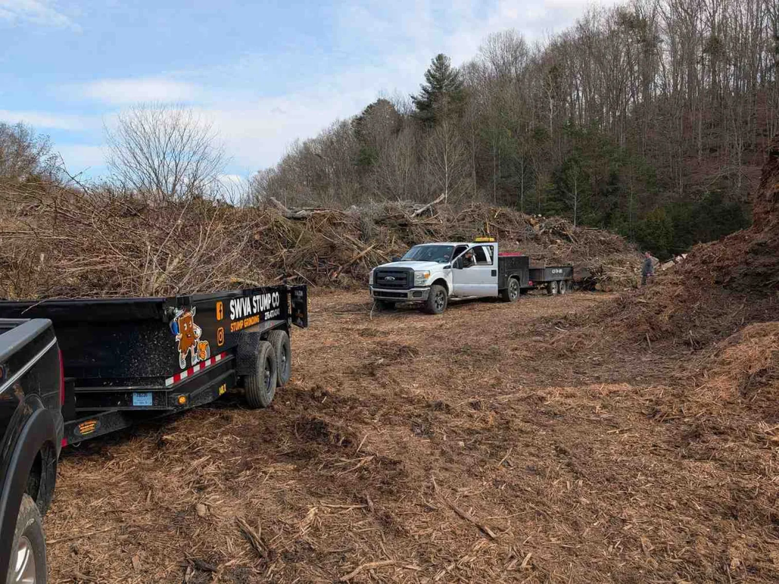 Work trucks with trailers at a tree clearing site covered in wood chips and debris near a forested hillside.