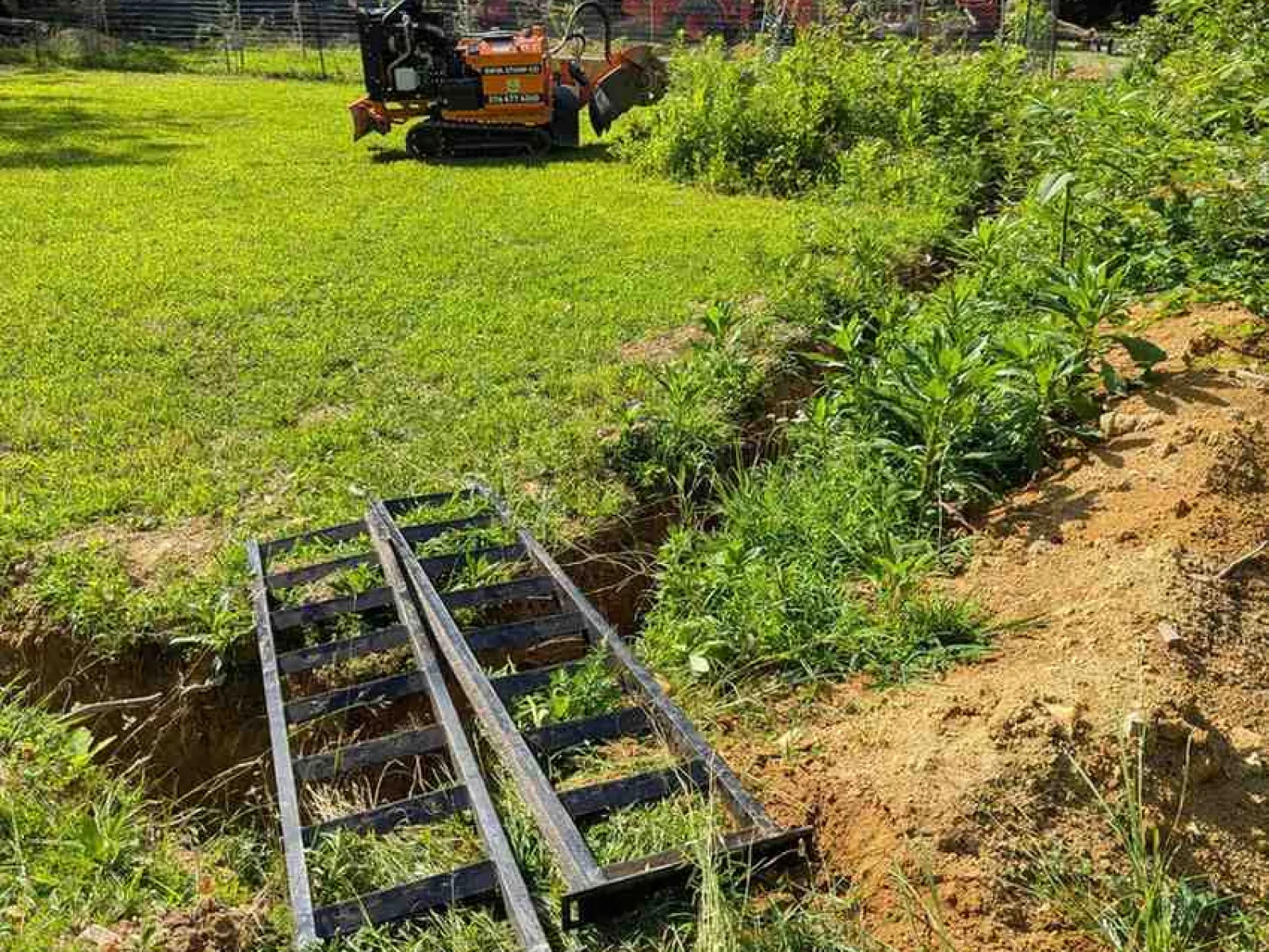 Metal ramps bridging a trench in a grassy yard with a small orange excavator and trees in the background.