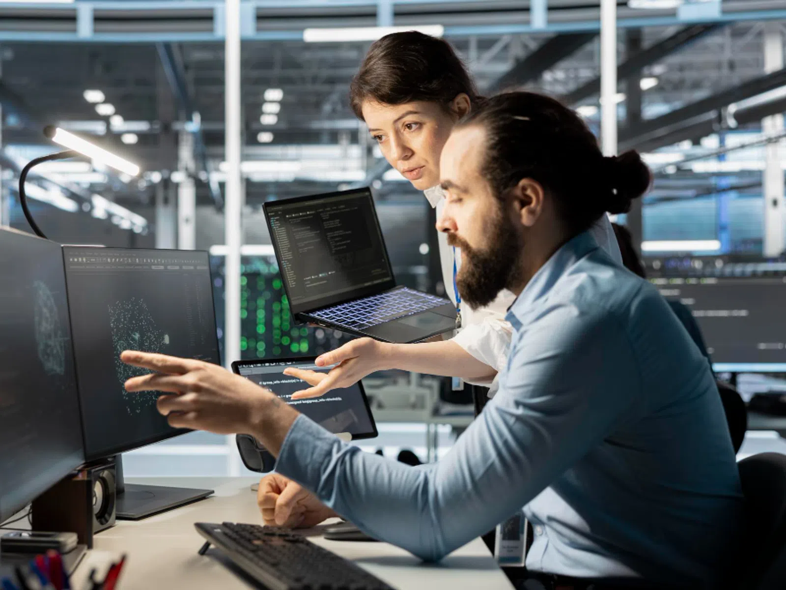 Two software developers collaborate while analyzing code and data on multiple computer screens in a high-tech office.
