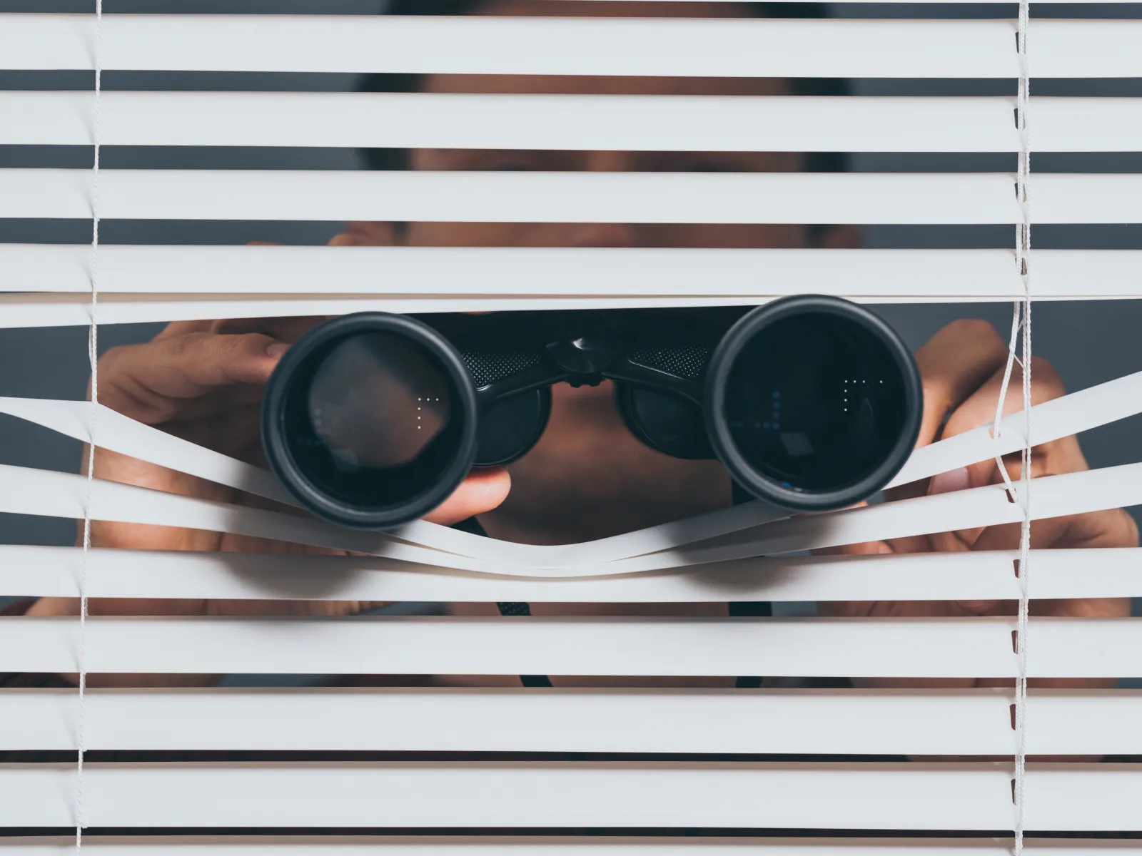 Person peeking through white blinds while holding black binoculars to observe outside discreetly.
