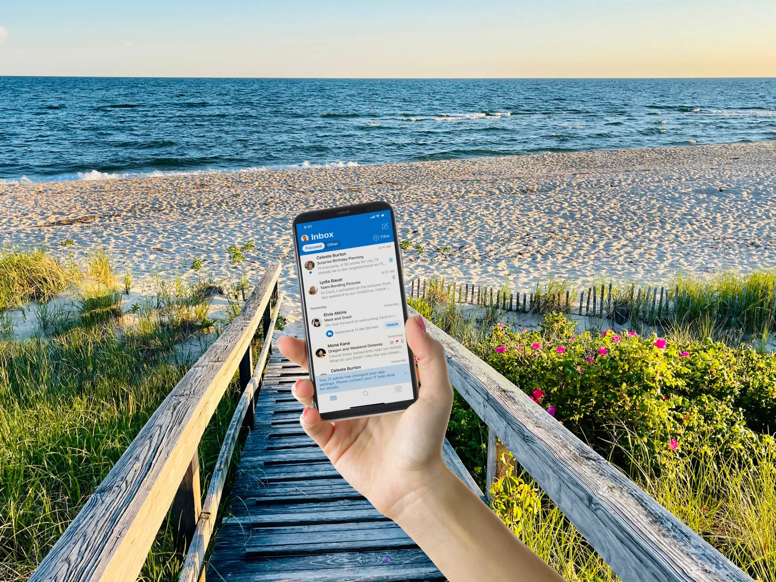 Person holding smartphone displaying email inbox on a wooden boardwalk by a sandy beach and ocean at sunset.