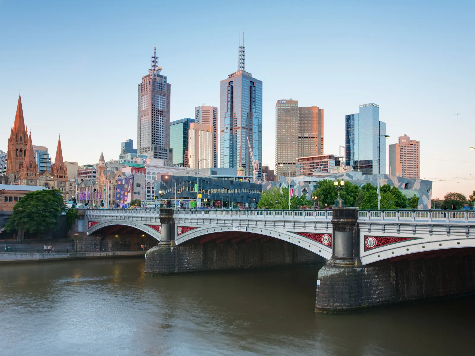 View of Melbourne city skyline with Yarra River and Princes Bridge at sunset under a clear sky.