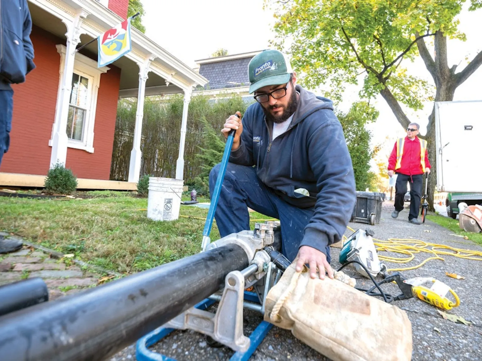 Plumber repairing a large pipe outdoors near a house on a sunny day with tools and equipment around.